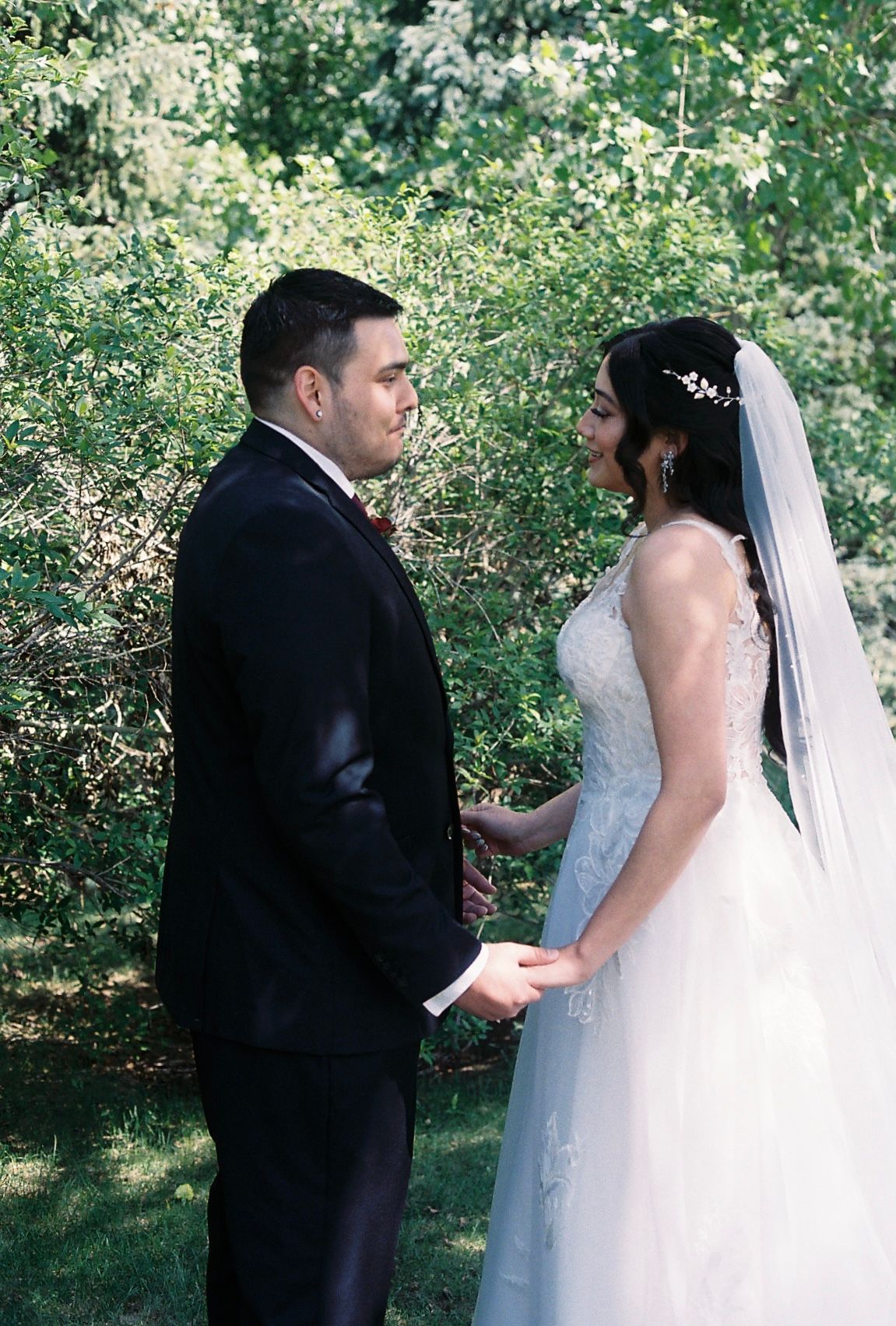 Bride and groom holding hands and gazing at each other lovingly in a sunlit outdoor garden setting.