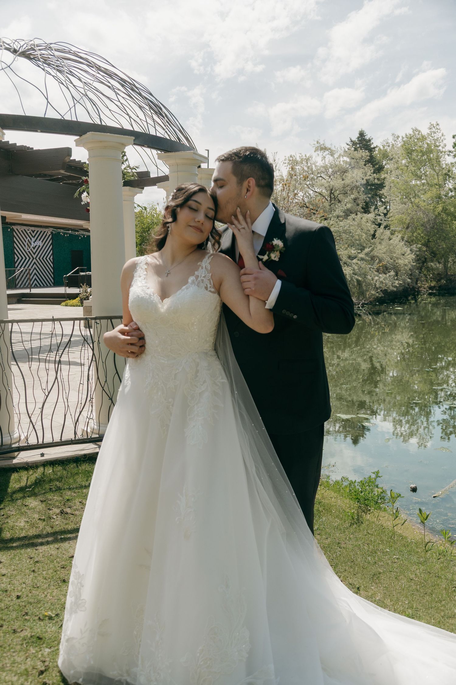 Groom in dark suit kisses bride in white lace gown near lakeside wedding pergola.