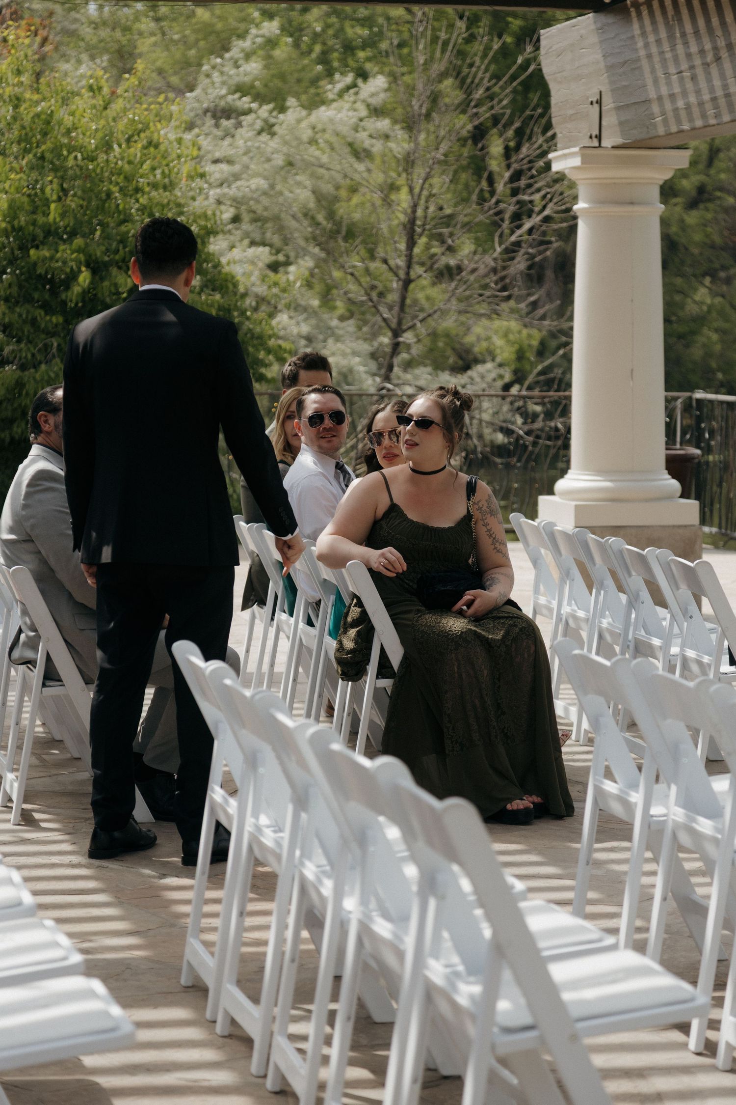 Wedding guests seated in white chairs at outdoor ceremony venue with stone columns and trees.