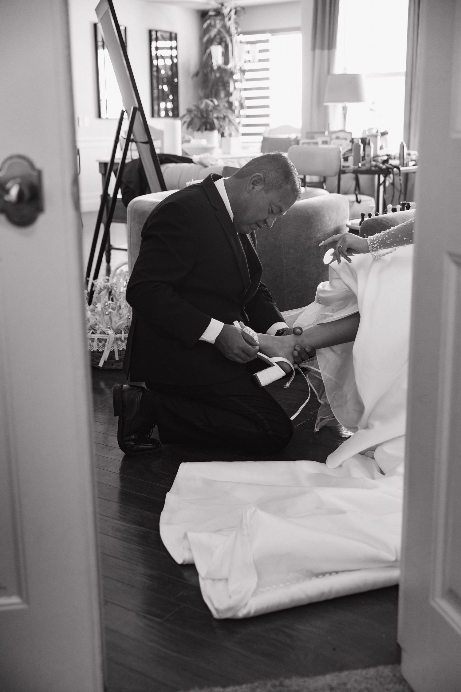 A groom kneels to help his bride with her shoes in a candid black and white wedding preparation photo.