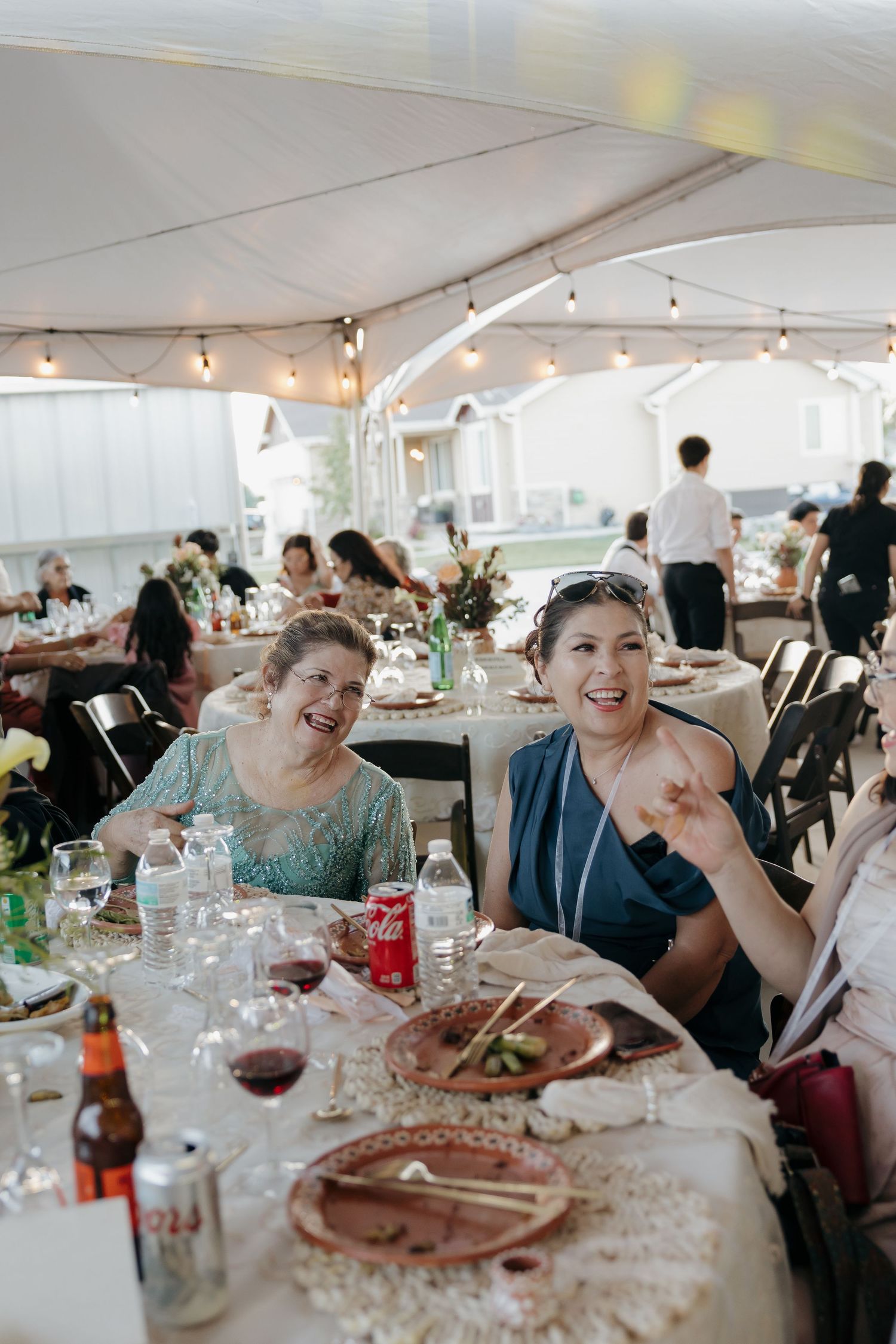 Two smiling women laugh and wave at camera during outdoor tented wedding reception dinner.