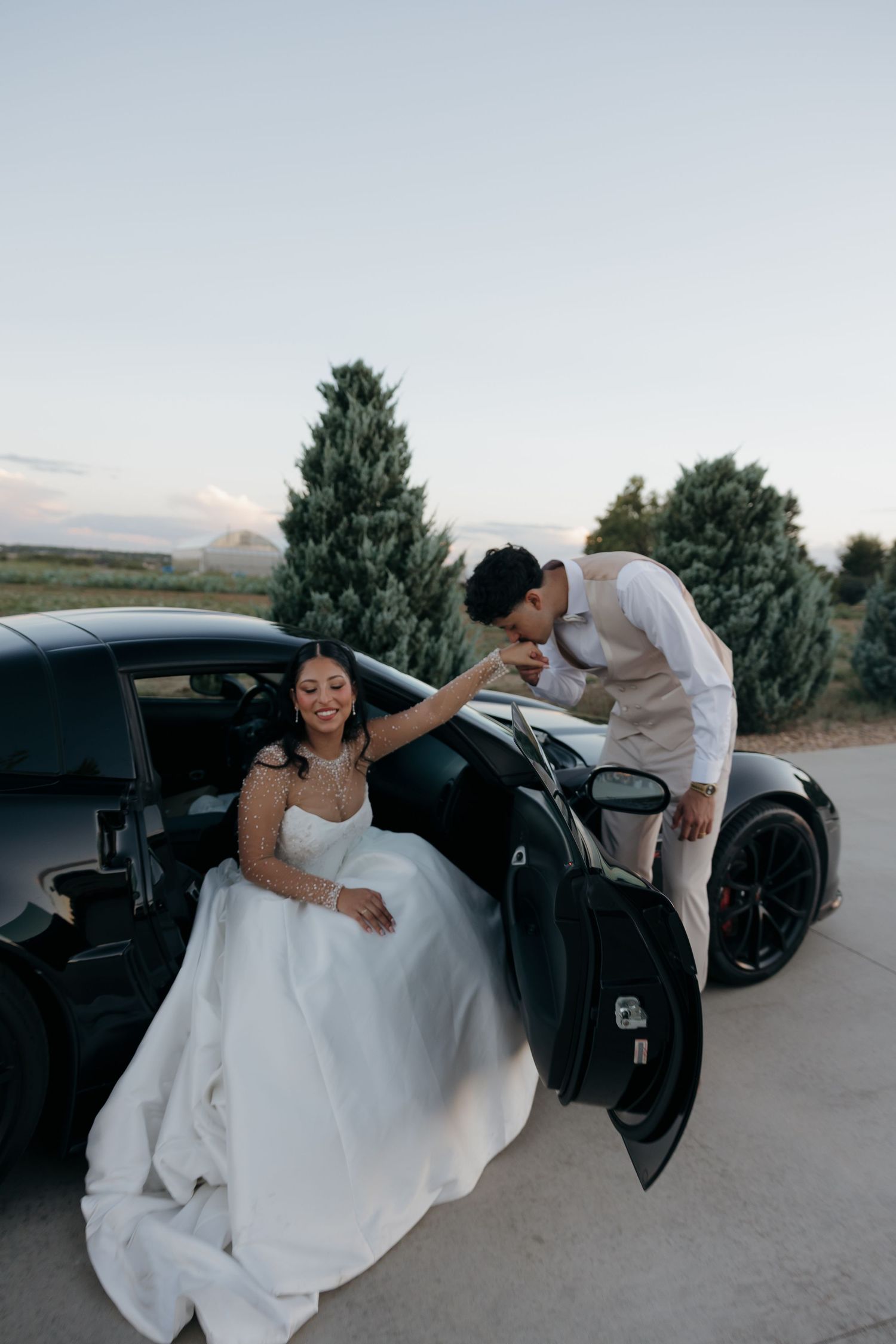 Groom kisses bride's hand as she sits in black sports car door at dusk wedding portrait.
