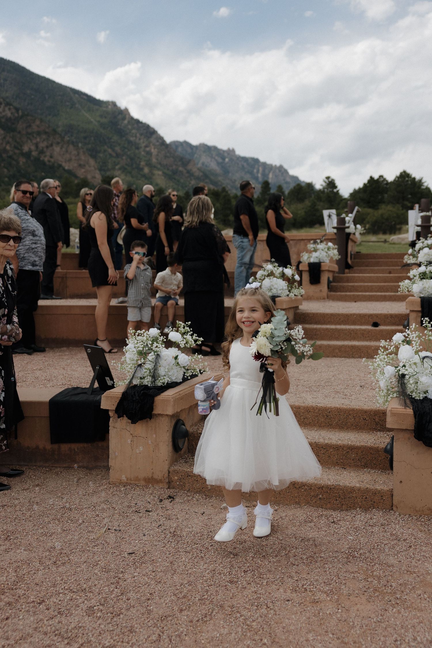 A flower girl in a white dress holds a bouquet at an outdoor mountain wedding ceremony with guests in black attire.
