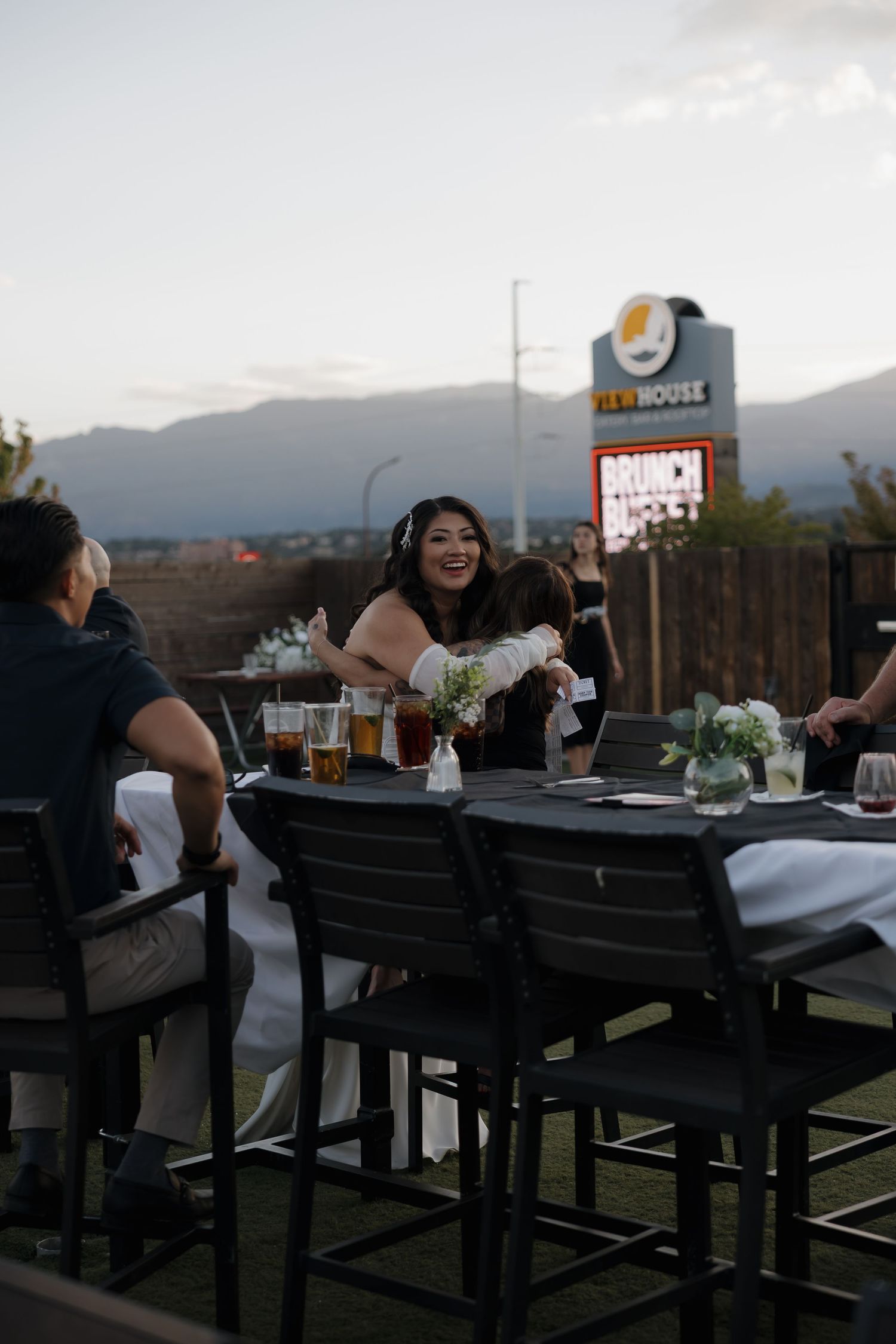 Smiling bride in wedding dress seated at outdoor patio table with guests at dusk reception.