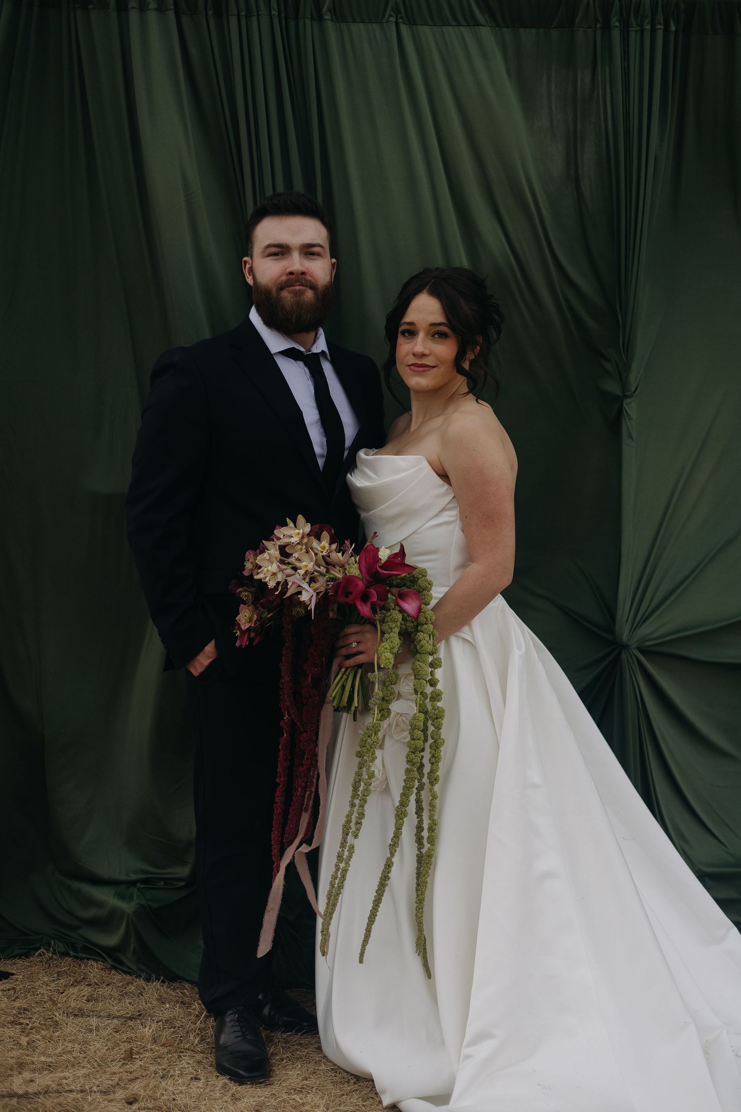 Wedding couple posing against dark green drapery with elegant floral arrangements.