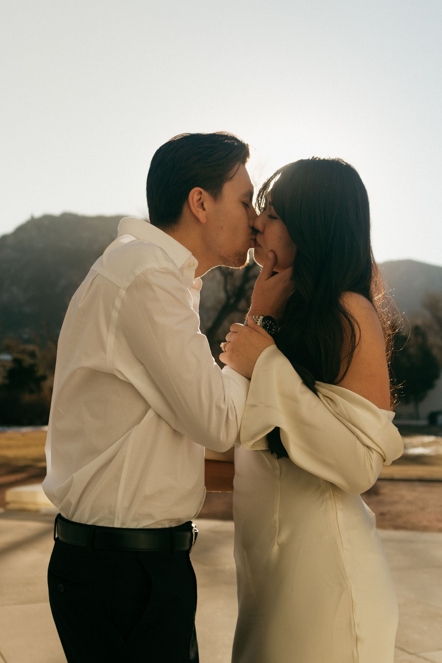 Couple sharing tender moments outdoors against a sunlit mountain landscape in formal attire.