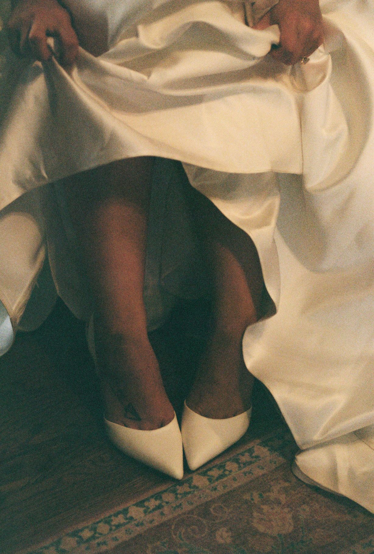 A pair of white heels worn with dark stockings against a cream-colored fabric backdrop and patterned carpet.