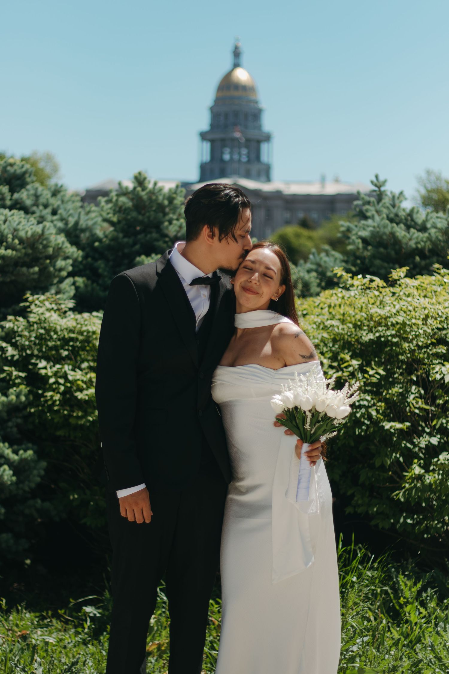 A couple shares romantic moments in front of a domed building on a sunny summer day with lush greenery.