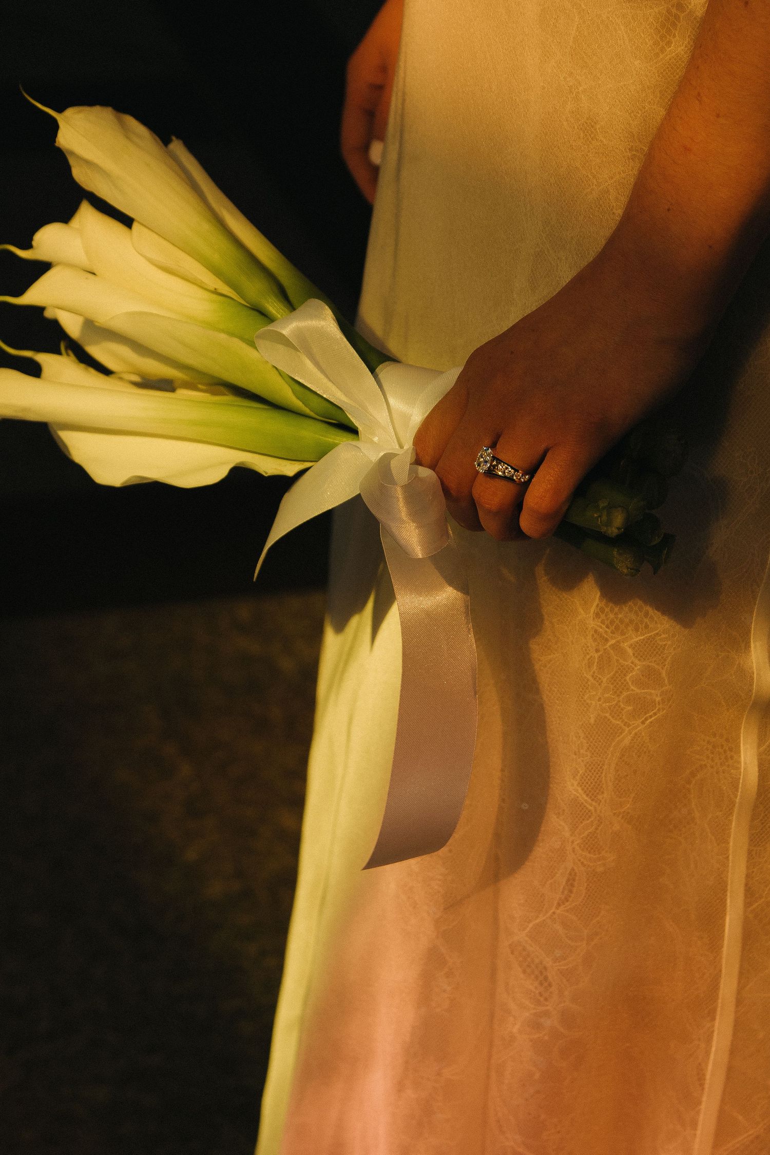 White calla lily bouquet held against lace wedding dress fabric.