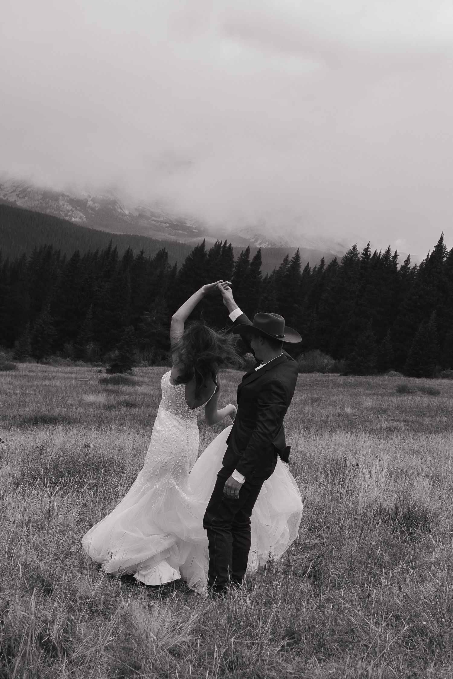 A couple dances in a mountain meadow with evergreen forests in the background.