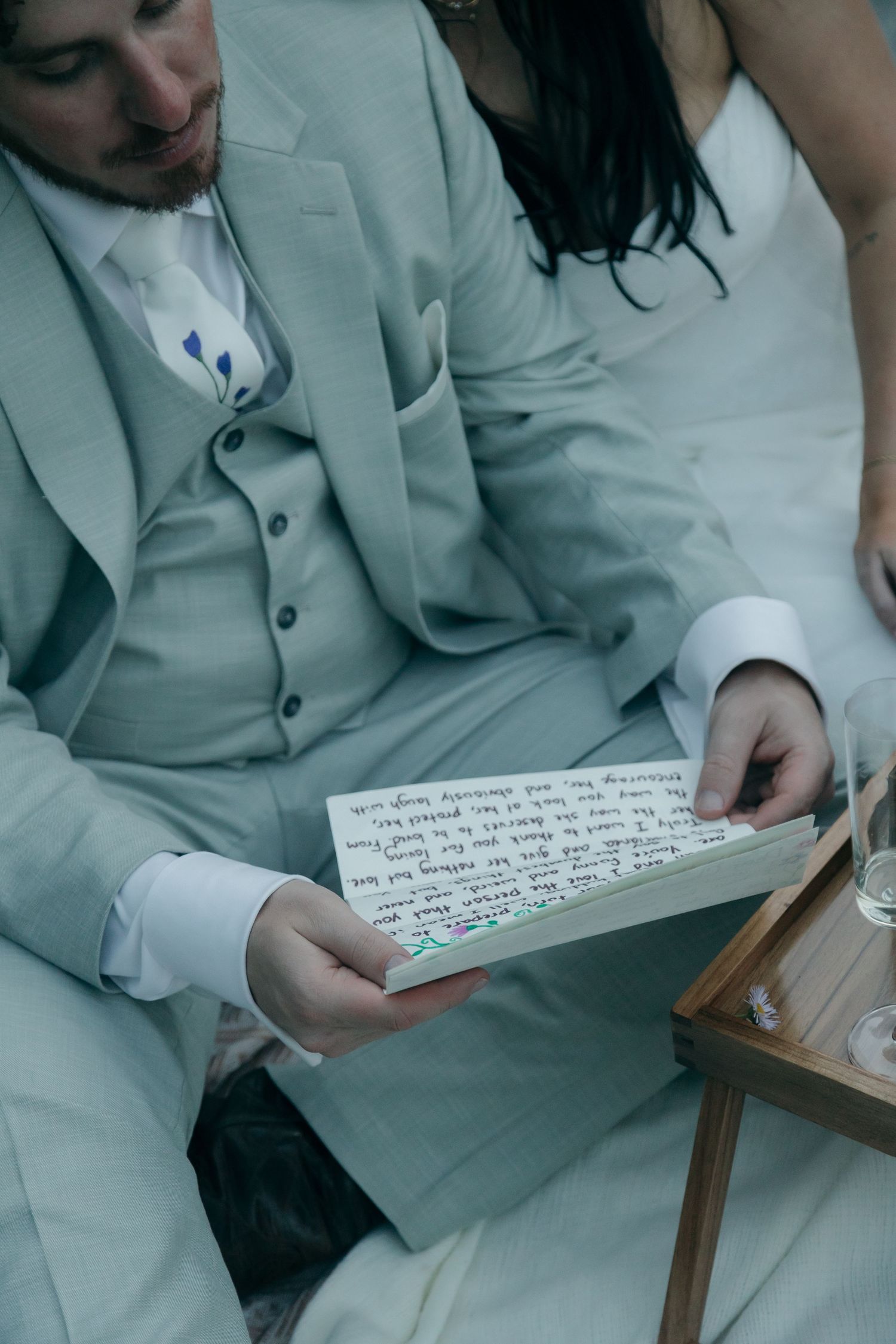 Groom in light gray suit reading handwritten wedding vow letter while seated beside bride during outdoor elopement picnic.