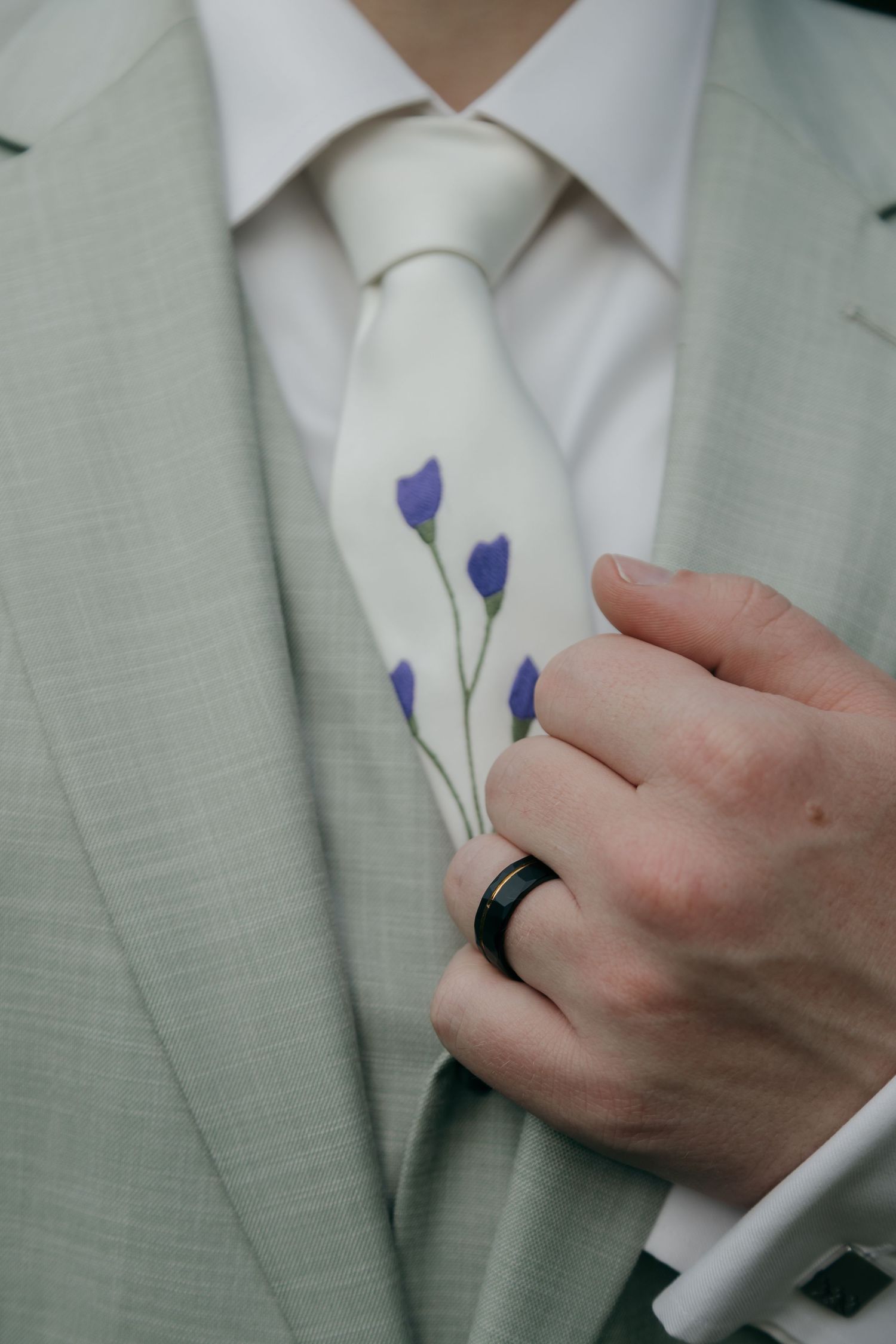 Groom in sage suit holds purple wildflowers against ivory tie, wearing black wedding band.