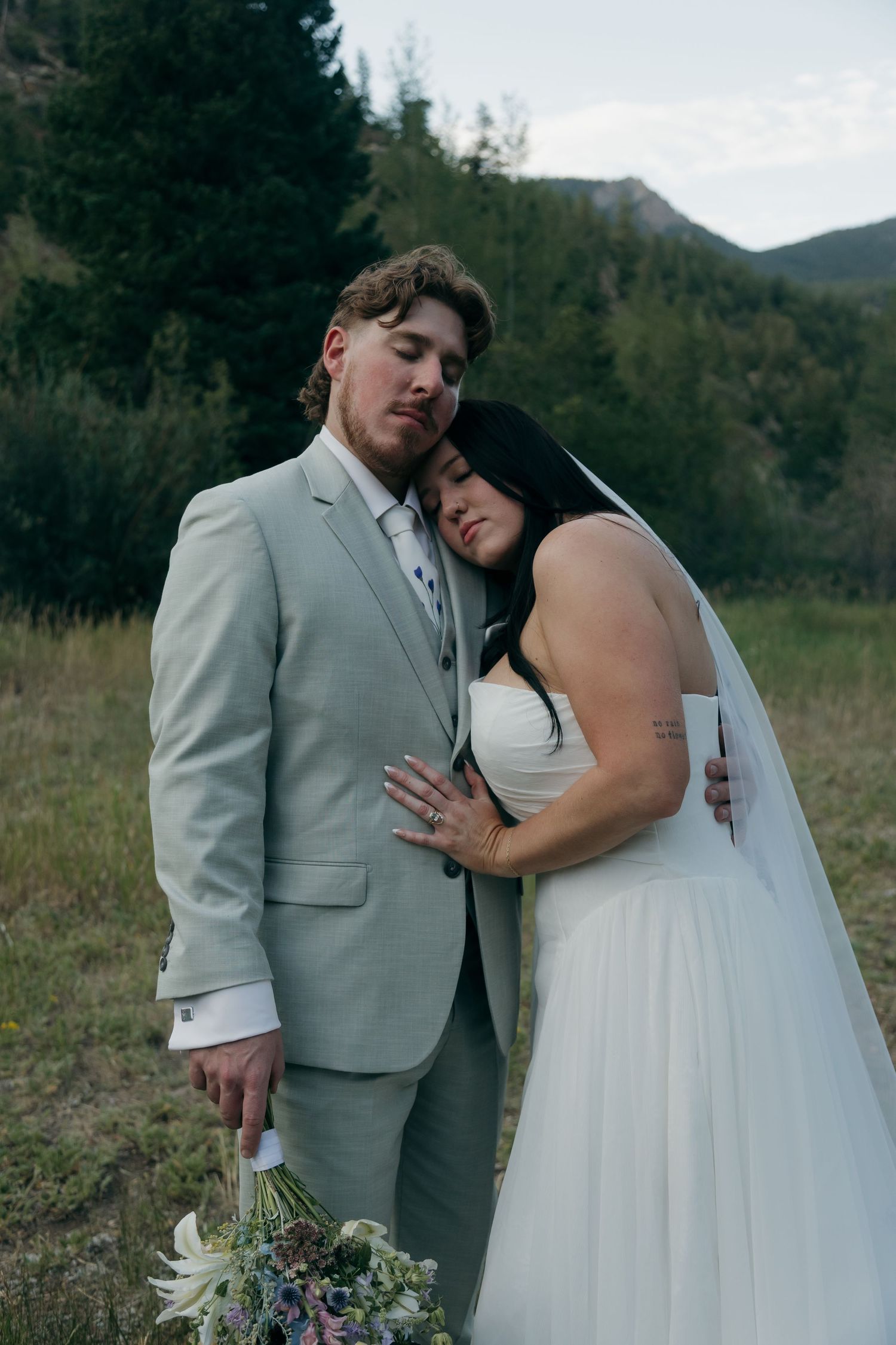 Bride leans her head on groom's shoulder as they pose together in a mountain meadow, holding a wildflower bouquet.