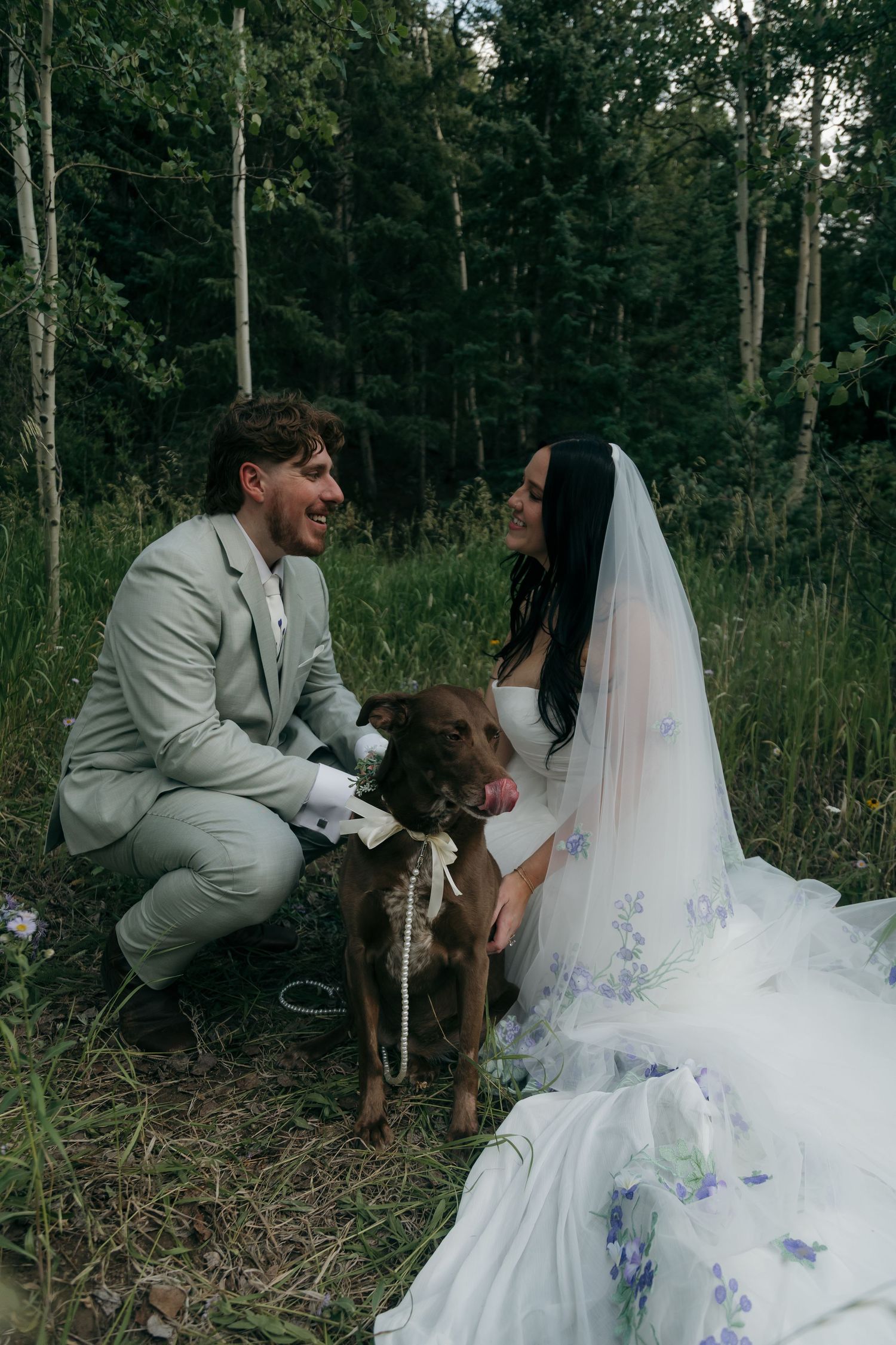 Bride and groom kneel with their brown dog in a forest during outdoor wedding celebration.