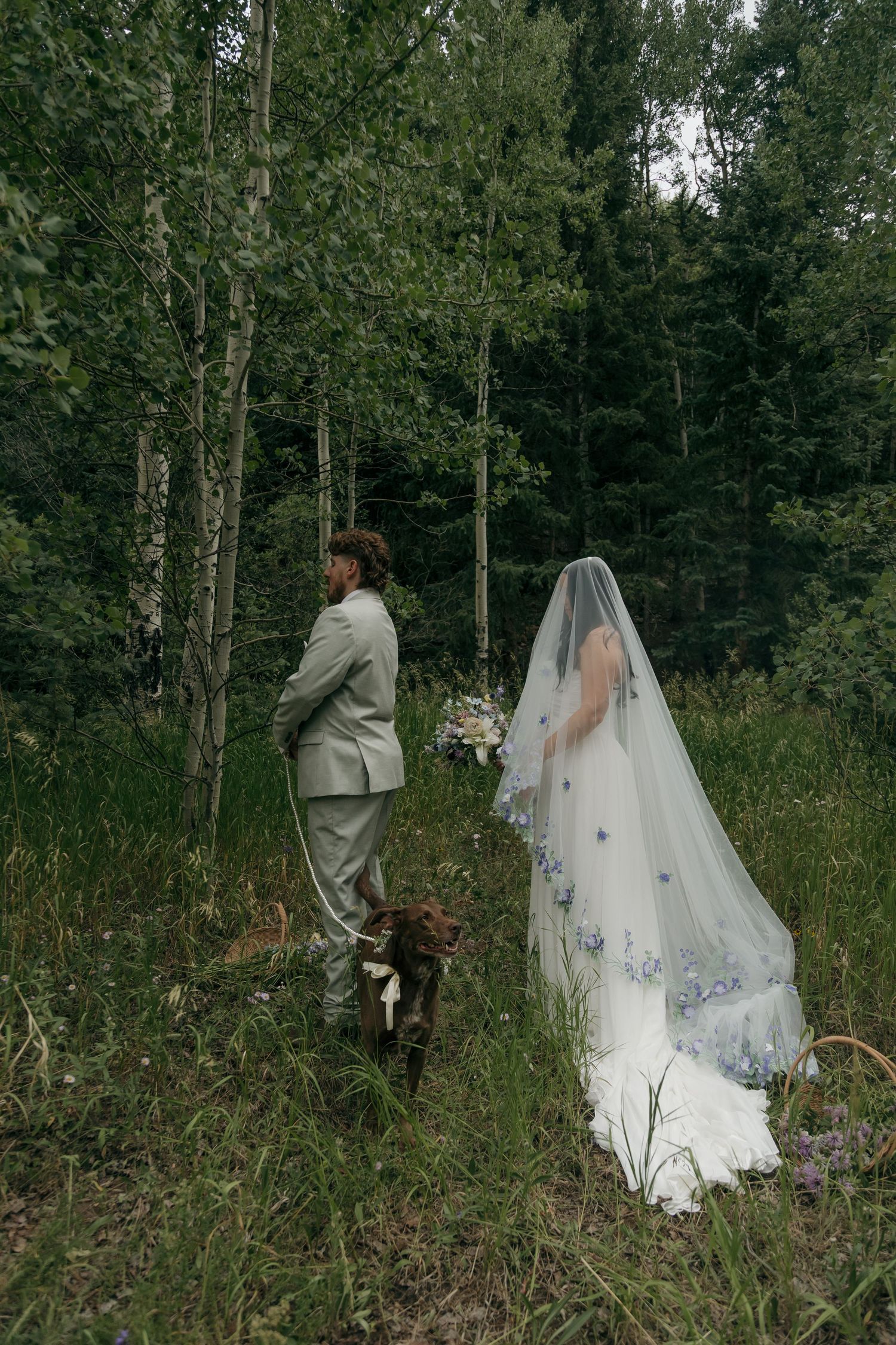 Bride in floral veil and groom in grey suit stand with dog in lush forest wedding photo.