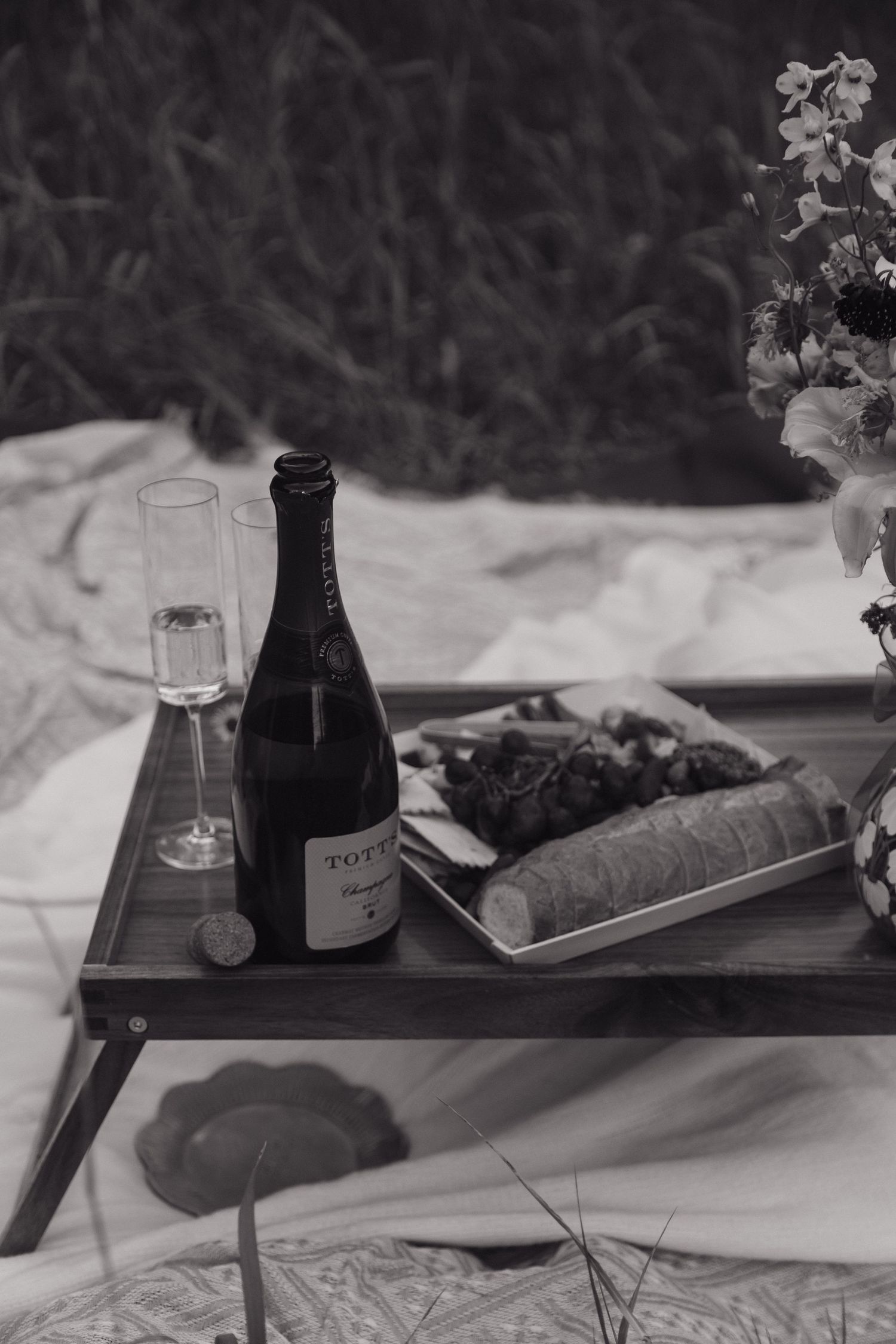 Black and white photo of champagne bottle, flute, and charcuterie board on rustic tray during outdoor wedding picnic.