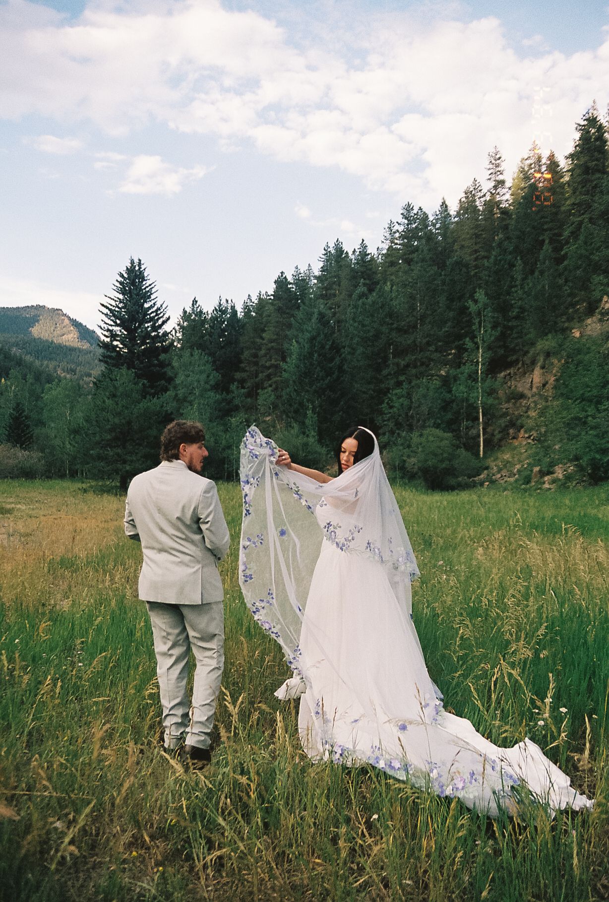 Bride lifts her floral embroidered veil while walking with groom through a scenic mountain meadow.
