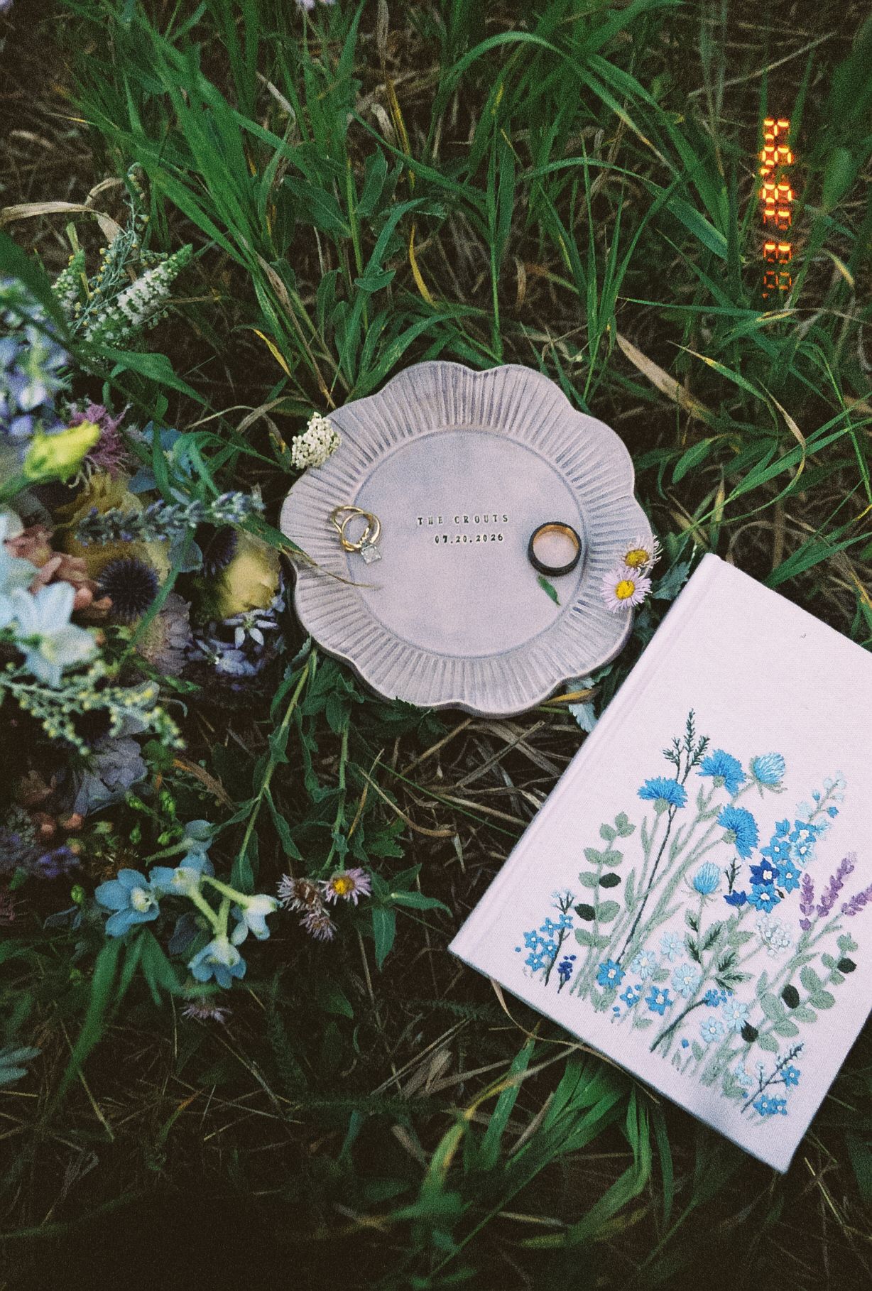 Wedding rings on ceramic dish beside wildflower bouquet and floral illustrated vow book in green grass.