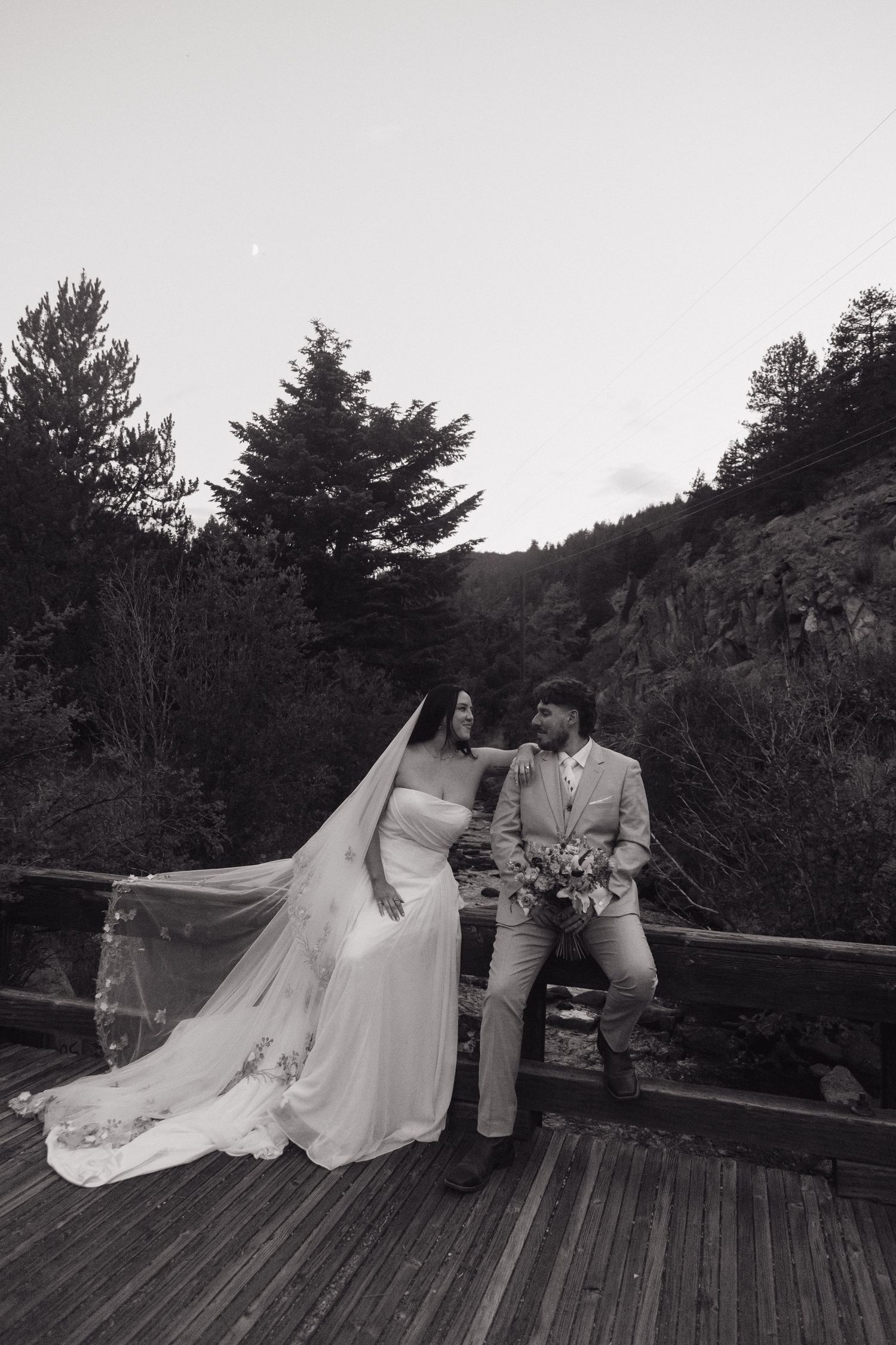 Black and white portrait of bride and groom on a rustic wooden bridge with flowing veil.