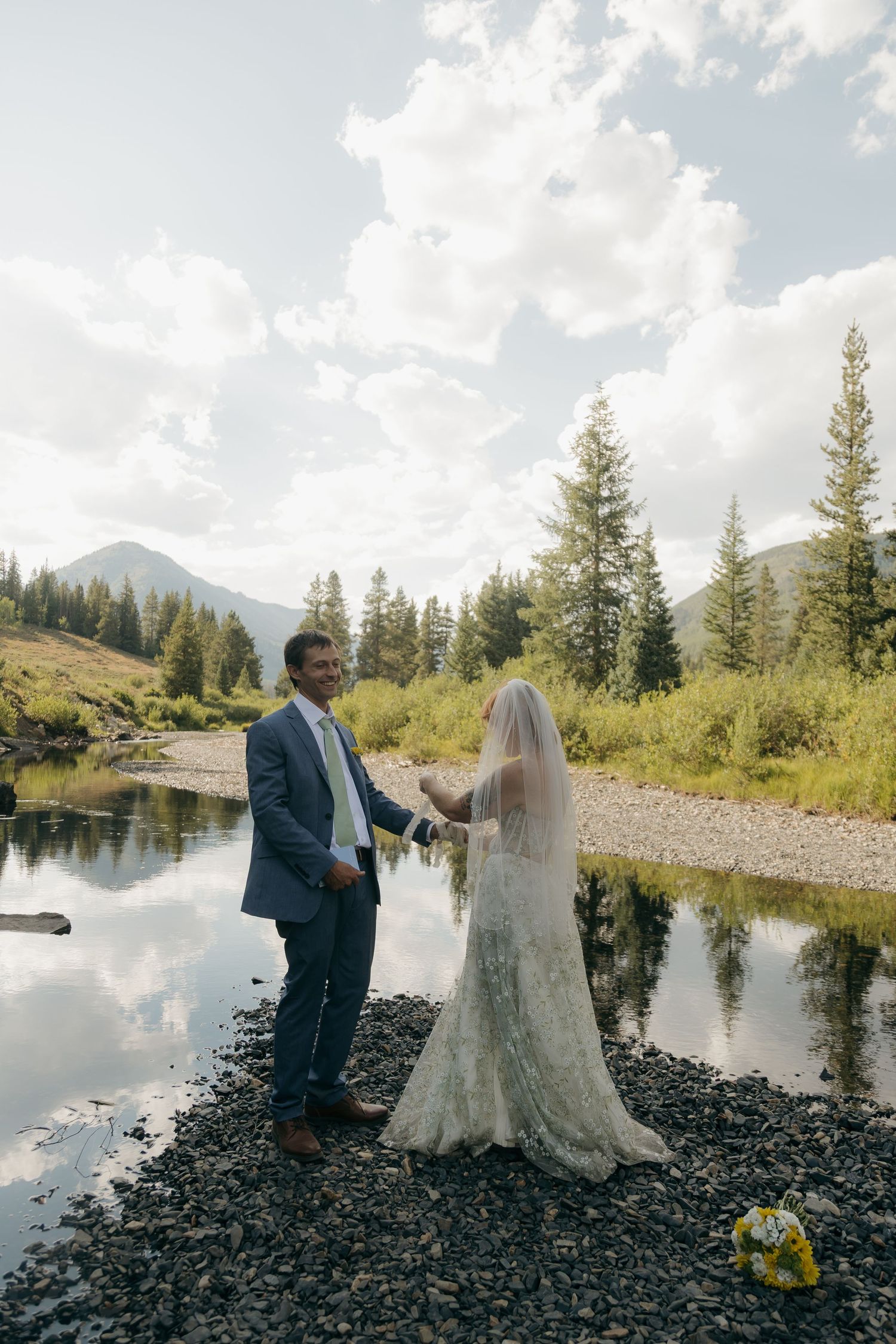 Bride and groom hold hands by a mountain river surrounded by pine trees during an outdoor wedding ceremony.