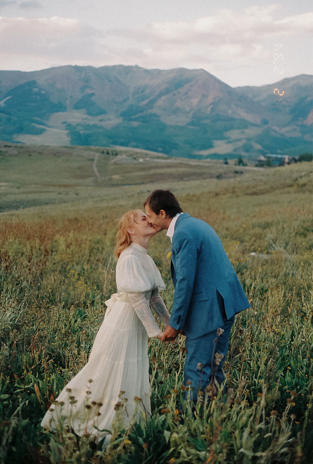 Newlyweds kissing in lush green mountain meadow with scenic rolling hills at golden hour.