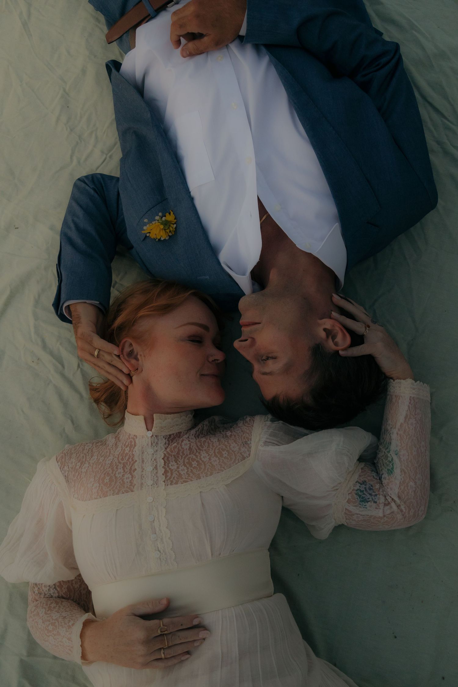 Bride and groom lying down face to face, she in white lace dress, he in blue suit with yellow flower boutonniere.