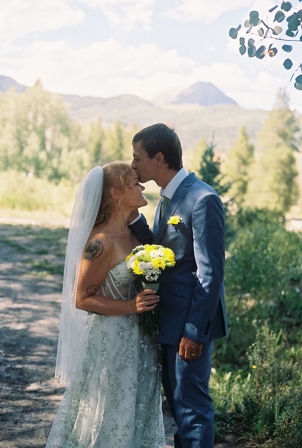 Bride and groom share first kiss holding yellow bouquet with mountain forest backdrop.