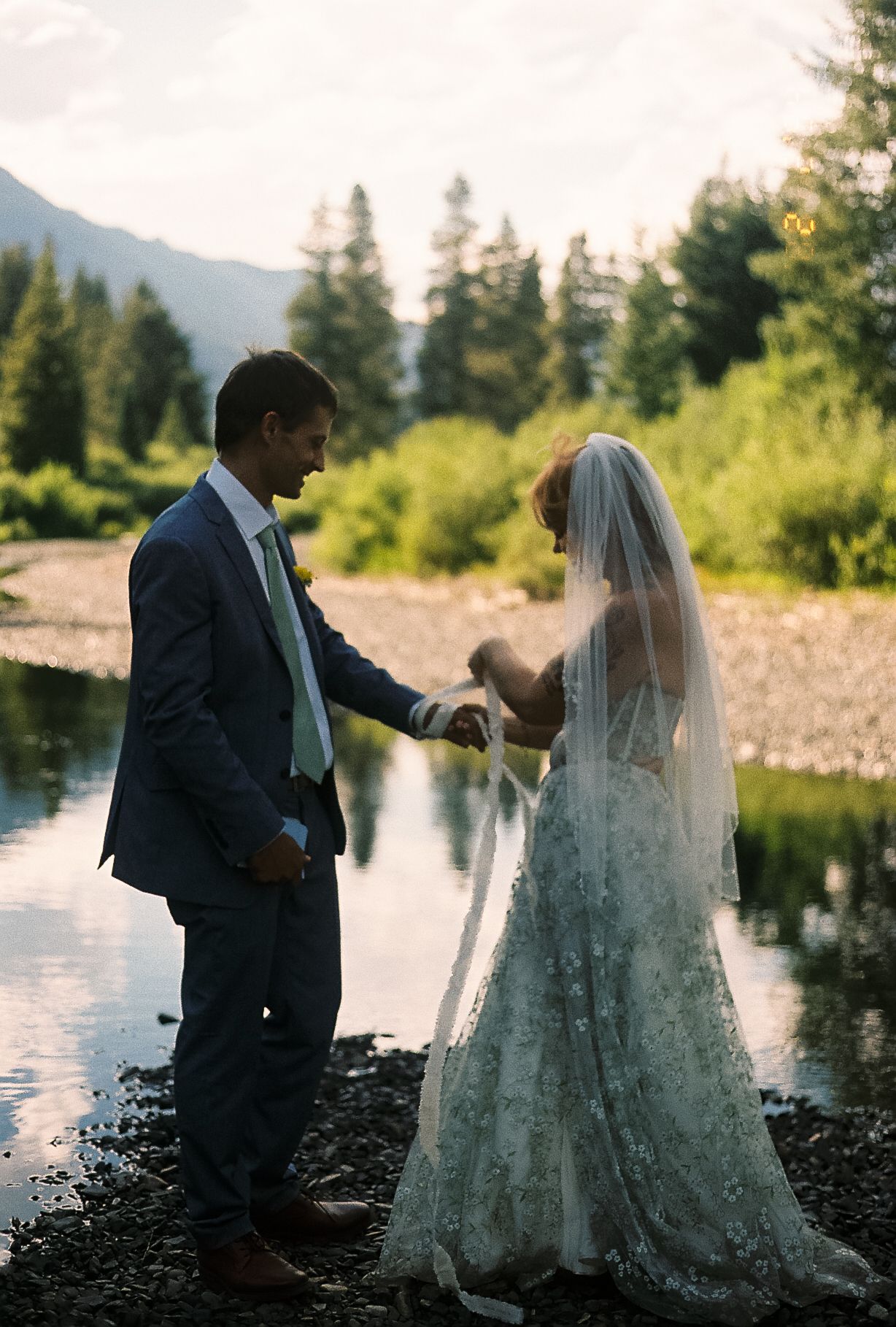 Bride in floral gown and veil holds hands with groom in blue suit by a mountain stream surrounded by lush greenery.