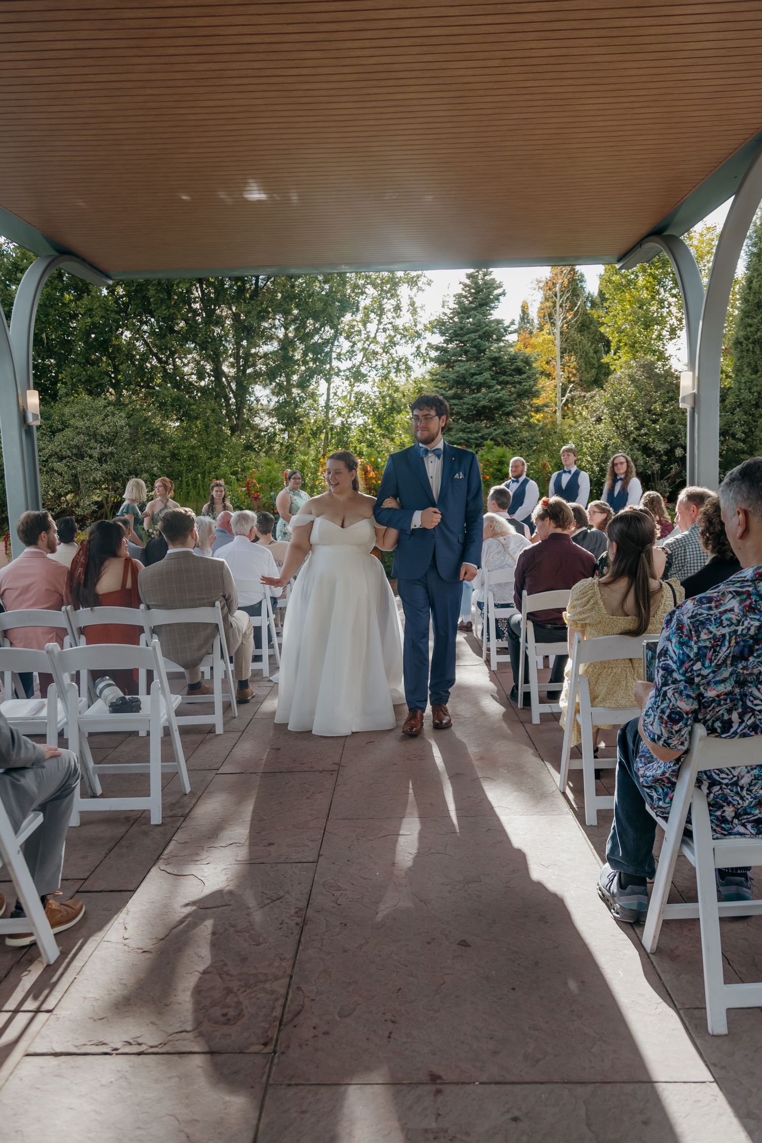 Newlyweds walk down the aisle at an outdoor garden wedding ceremony with white chairs and lush trees.