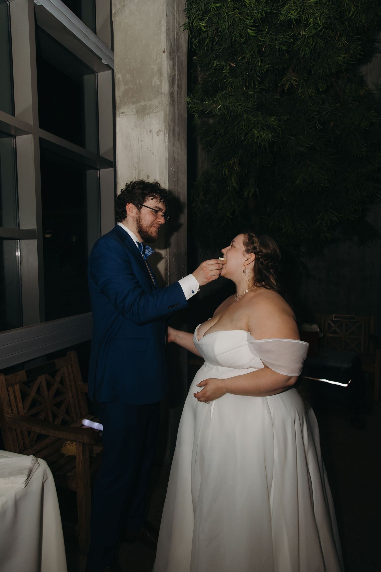 Groom in blue suit feeds bride wedding cake during outdoor evening wedding reception.