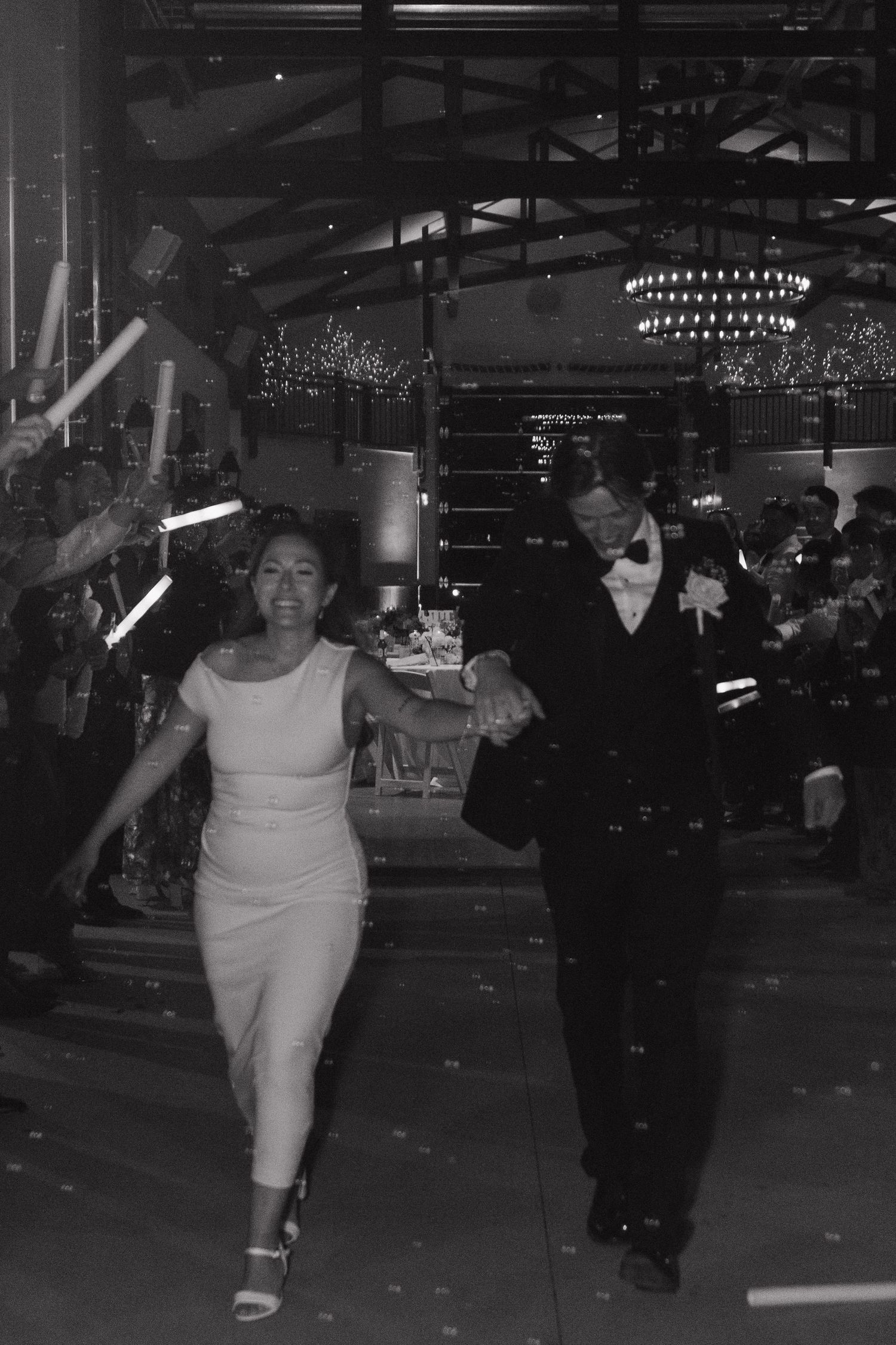 A black and white photo captures a couple dancing together under elegant chandeliers at an indoor venue.