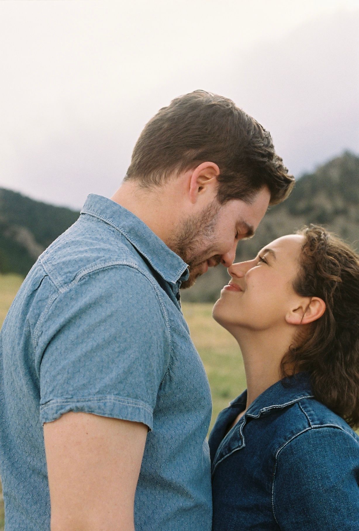 A romantic couple sharing an intimate moment outdoors with mountains in the background, both wearing denim shirts.