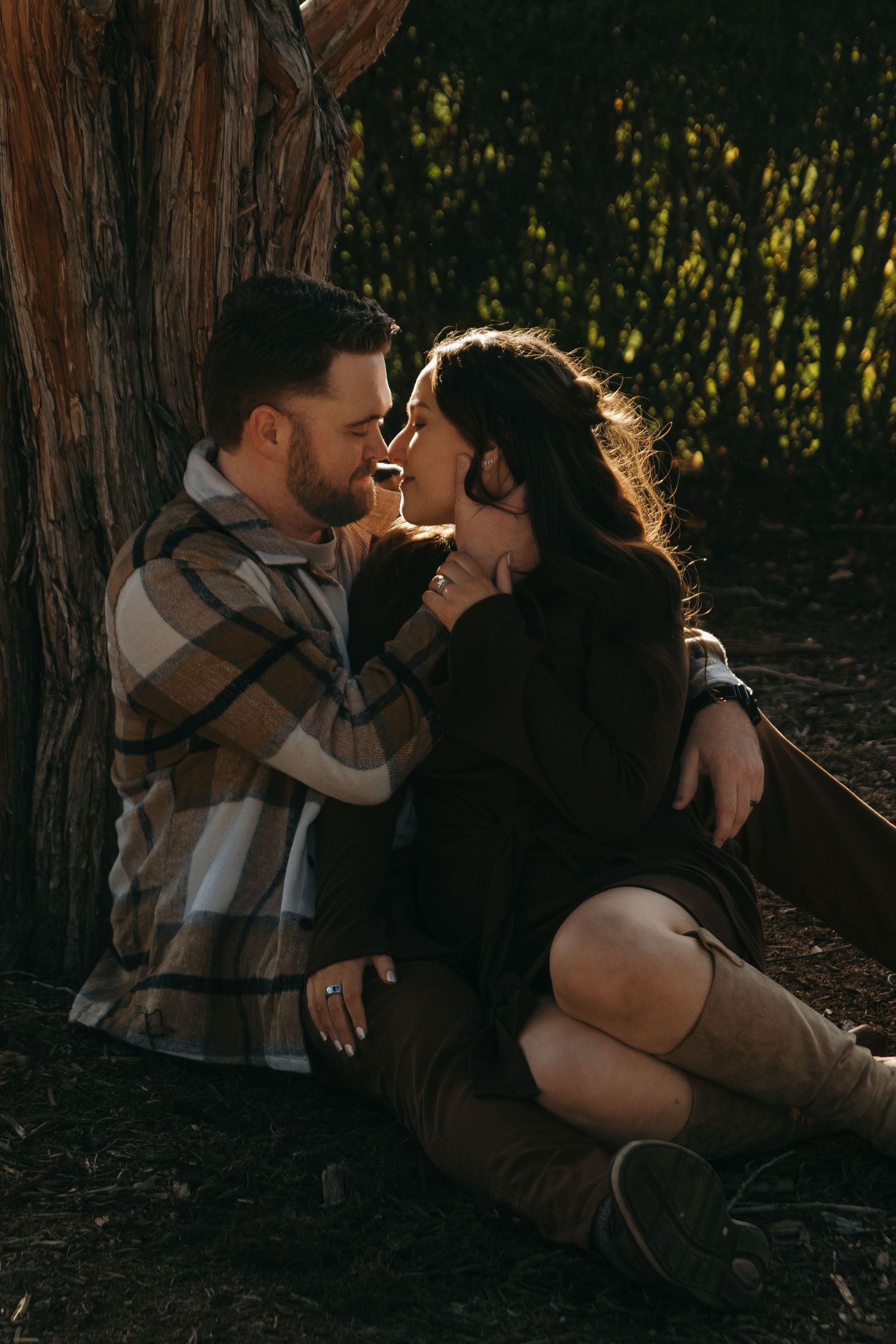 Couple sharing a romantic moment leaning against a tree in golden hour light, woman in brown boots and black dress.