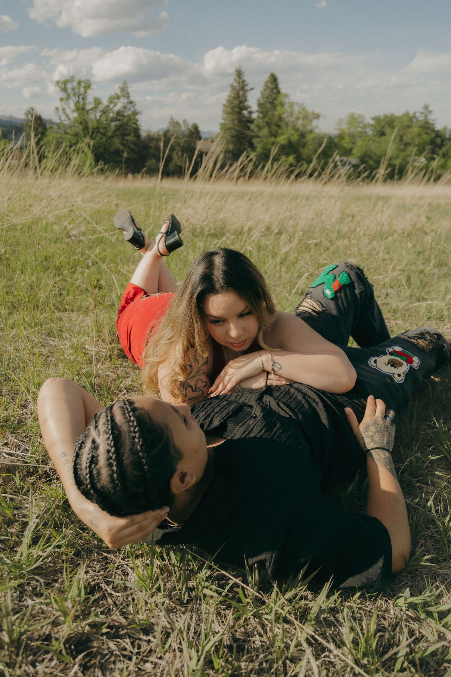 Couple relaxing in a grassy field, woman with curly hair resting on man with braids on a sunny day.