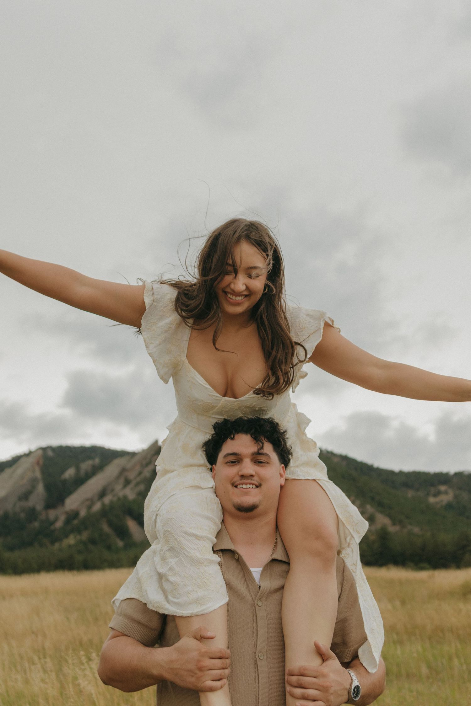 Smiling woman sitting on mans shoulders with arms outstretched in a golden field with mountains.