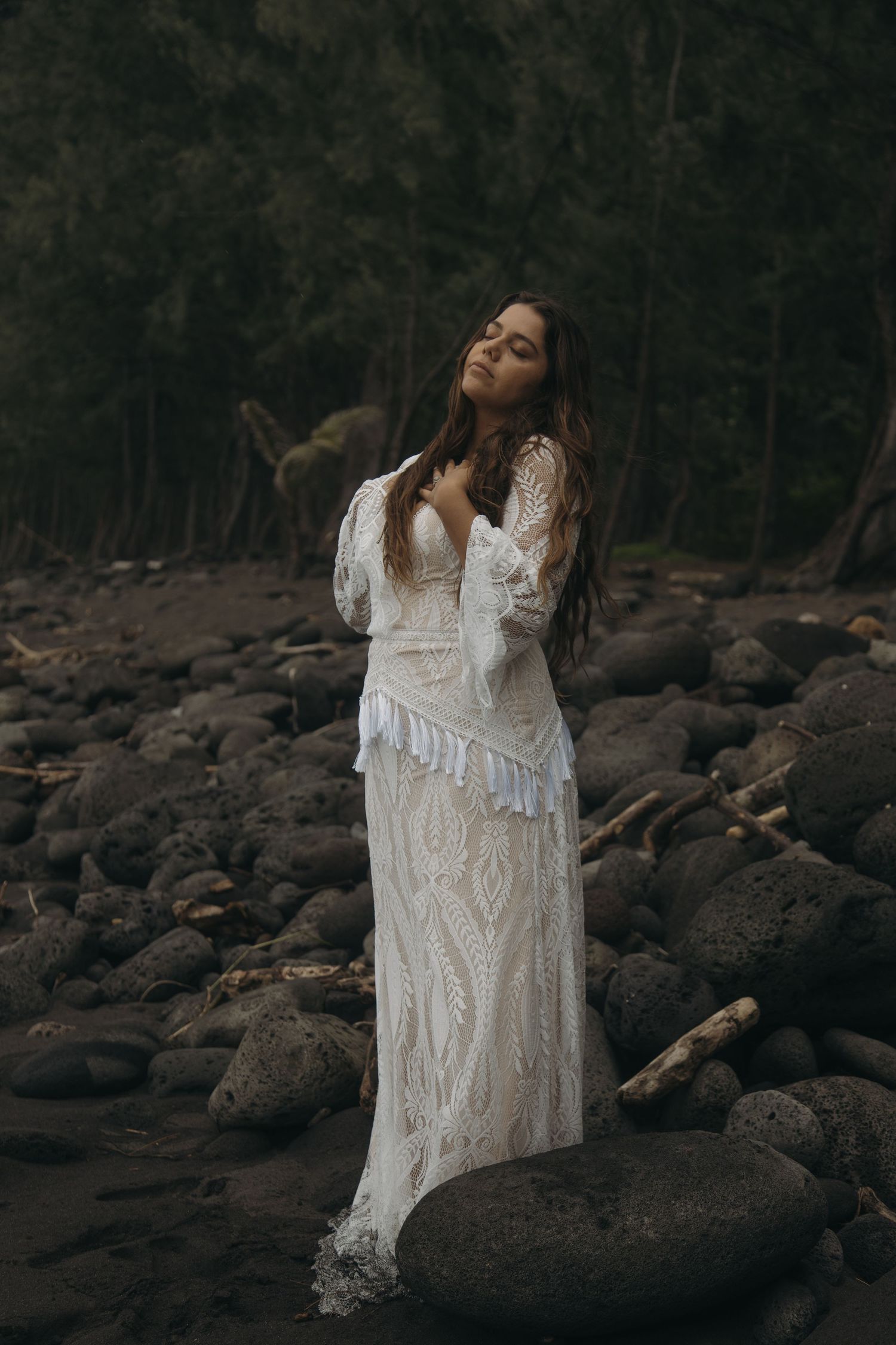 Bride in bohemian lace wedding gown standing gracefully on rocky shoreline surrounded by dark forest.