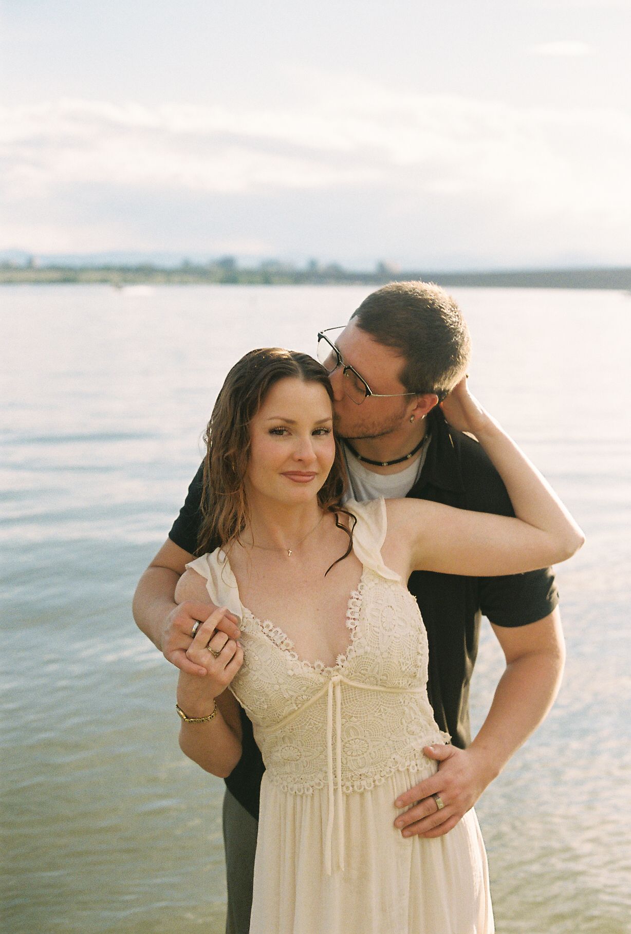 A man kisses a woman on the cheek while embracing her from behind near a calm lake during golden hour.