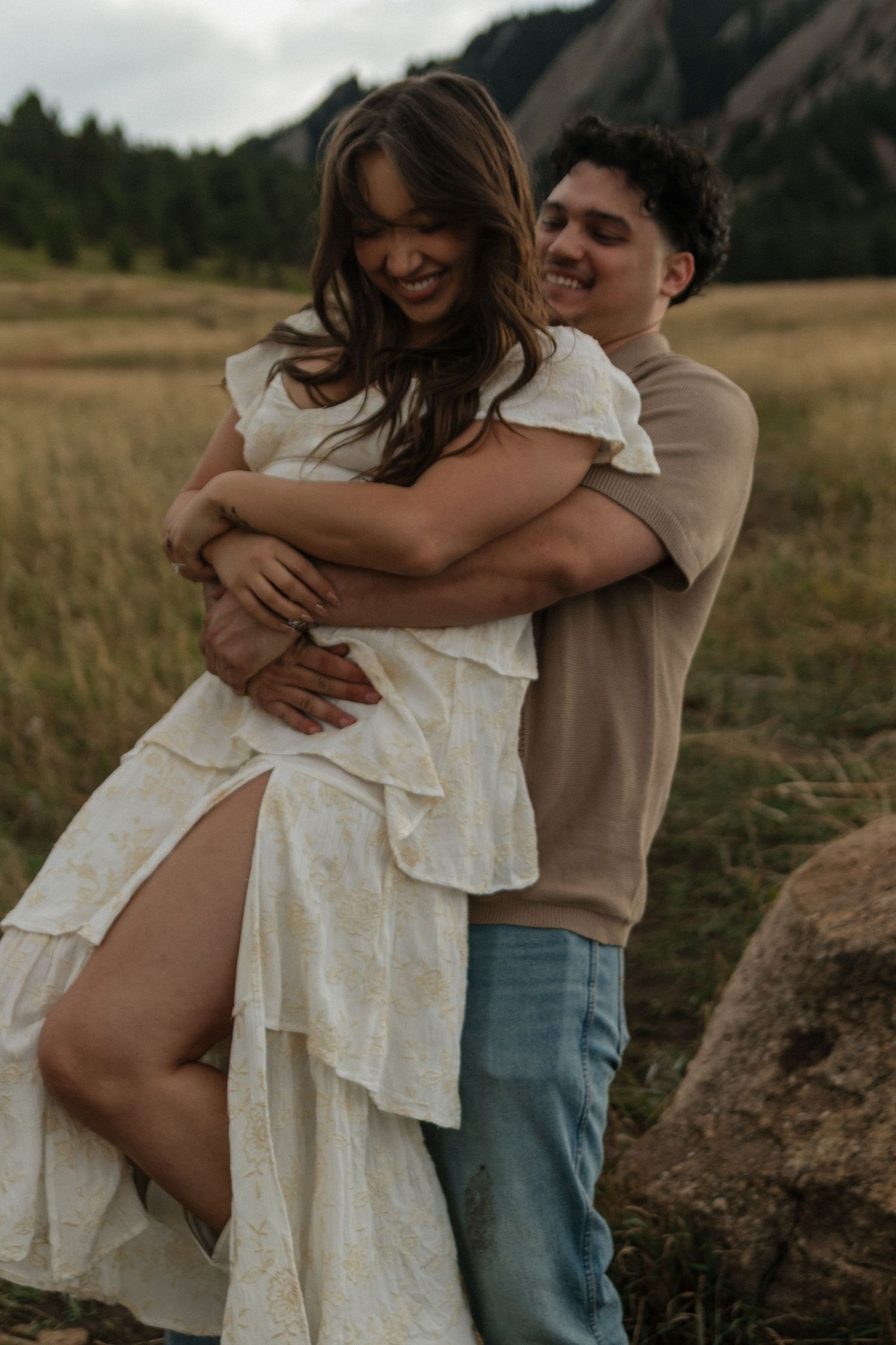 Man hugging woman in white ruffled dress from behind, both smiling in a grassy mountain field.