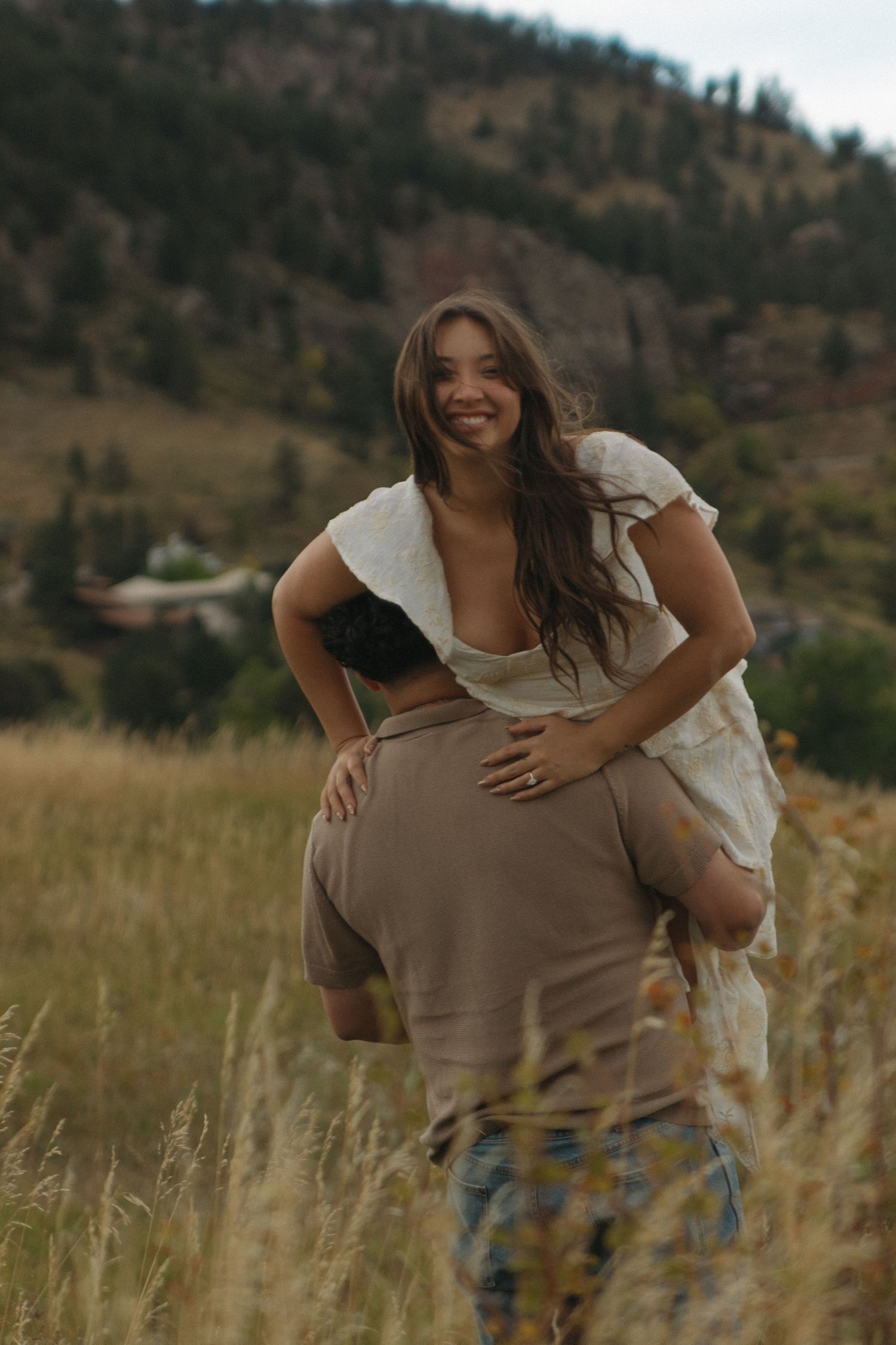 Man giving smiling woman in white dress a piggyback ride through golden mountain meadow.