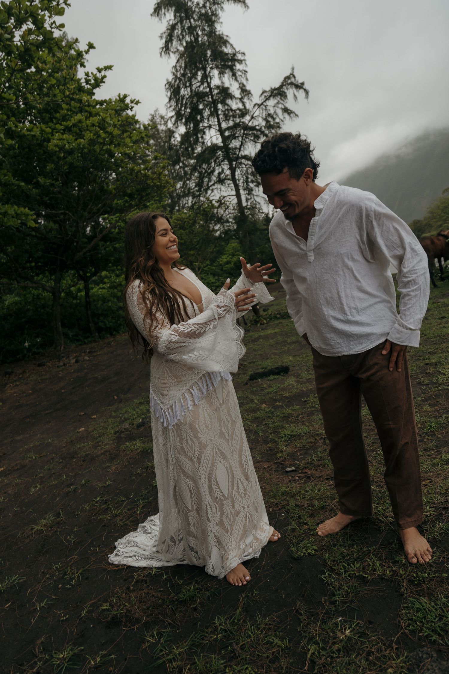 Couple in bohemian wedding attire sharing a joyful moment outdoors amid lush green trees and misty mountains.