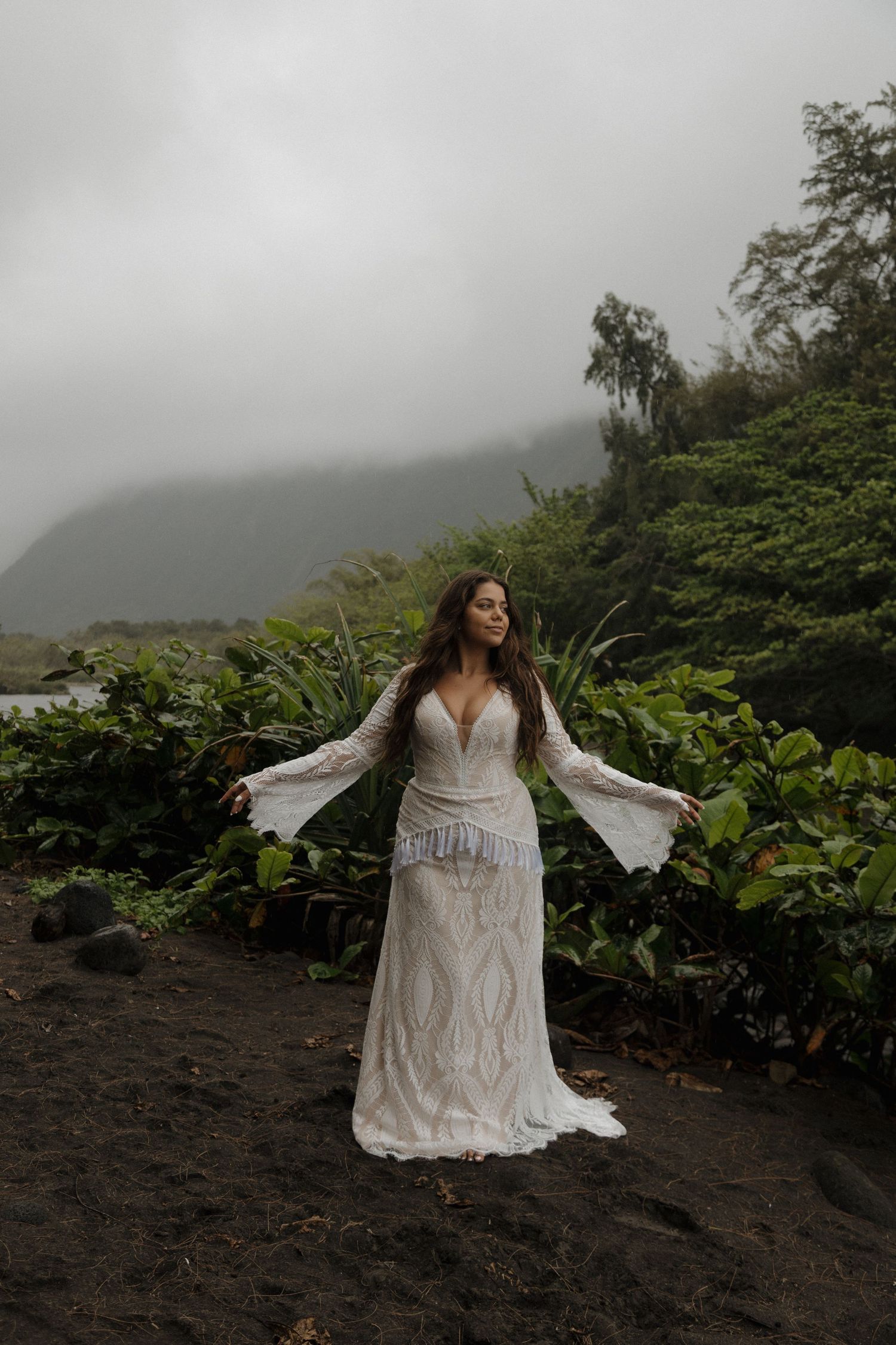Bride in boho lace gown with bell sleeves stands on black sand beach with misty mountains.