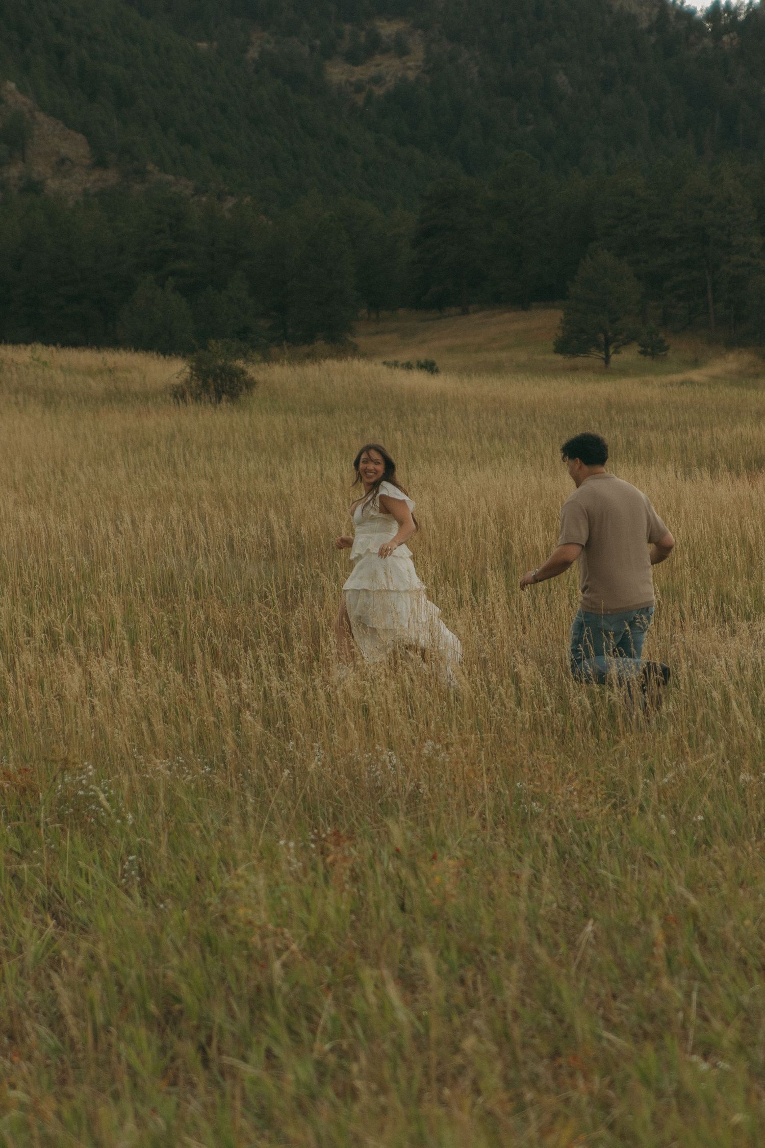 Couple running through golden meadow with pine forest backdrop during outdoor engagement session.