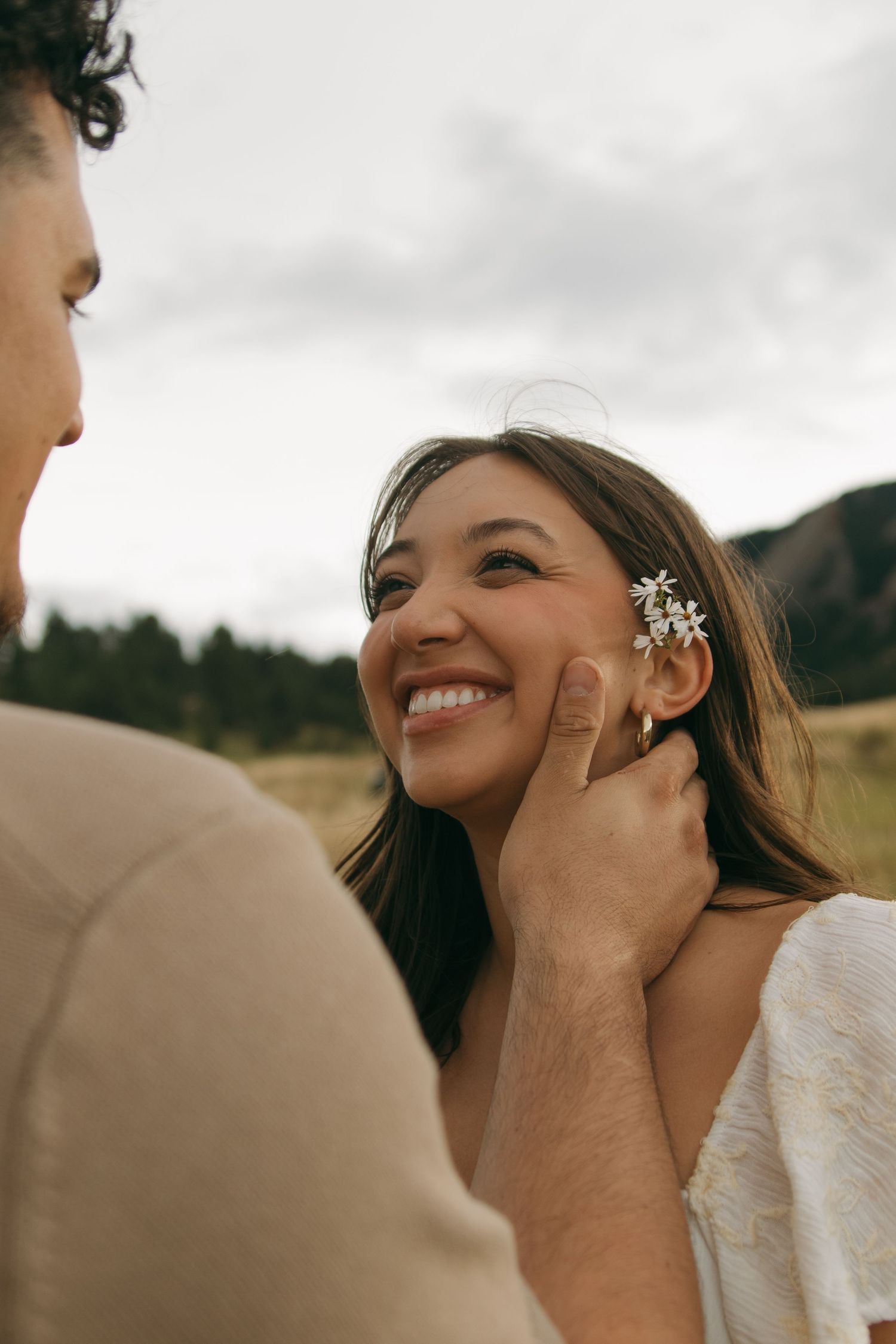 Smiling bride with flower hair accessory gazing up at groom outdoors in mountain field.