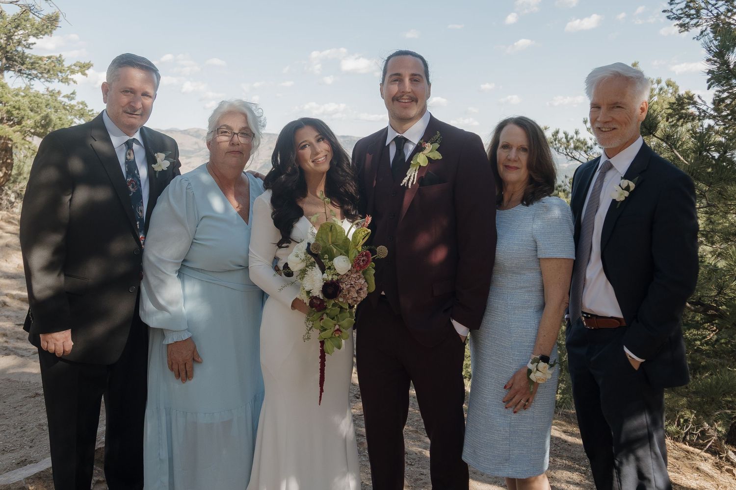 Wedding party of six posing outdoors in formal attire with bride in white gown and groom in burgundy suit.