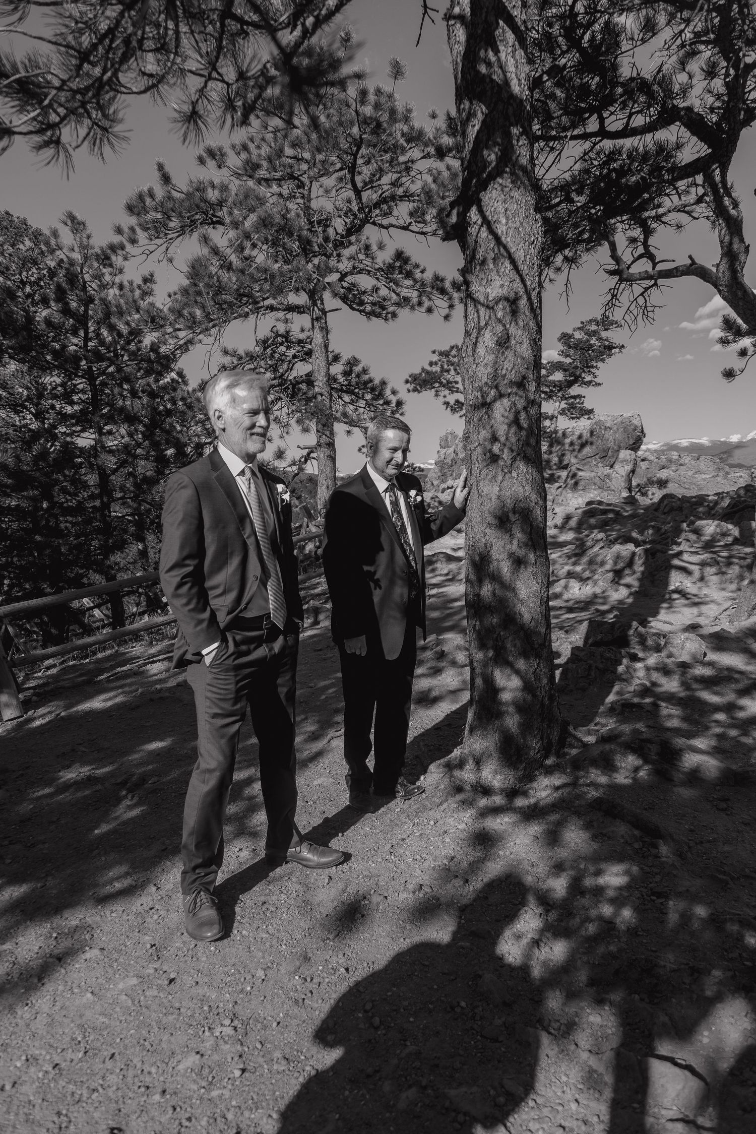 Black and white photo of two suited men conversing among pine trees at an outdoor mountain wedding.