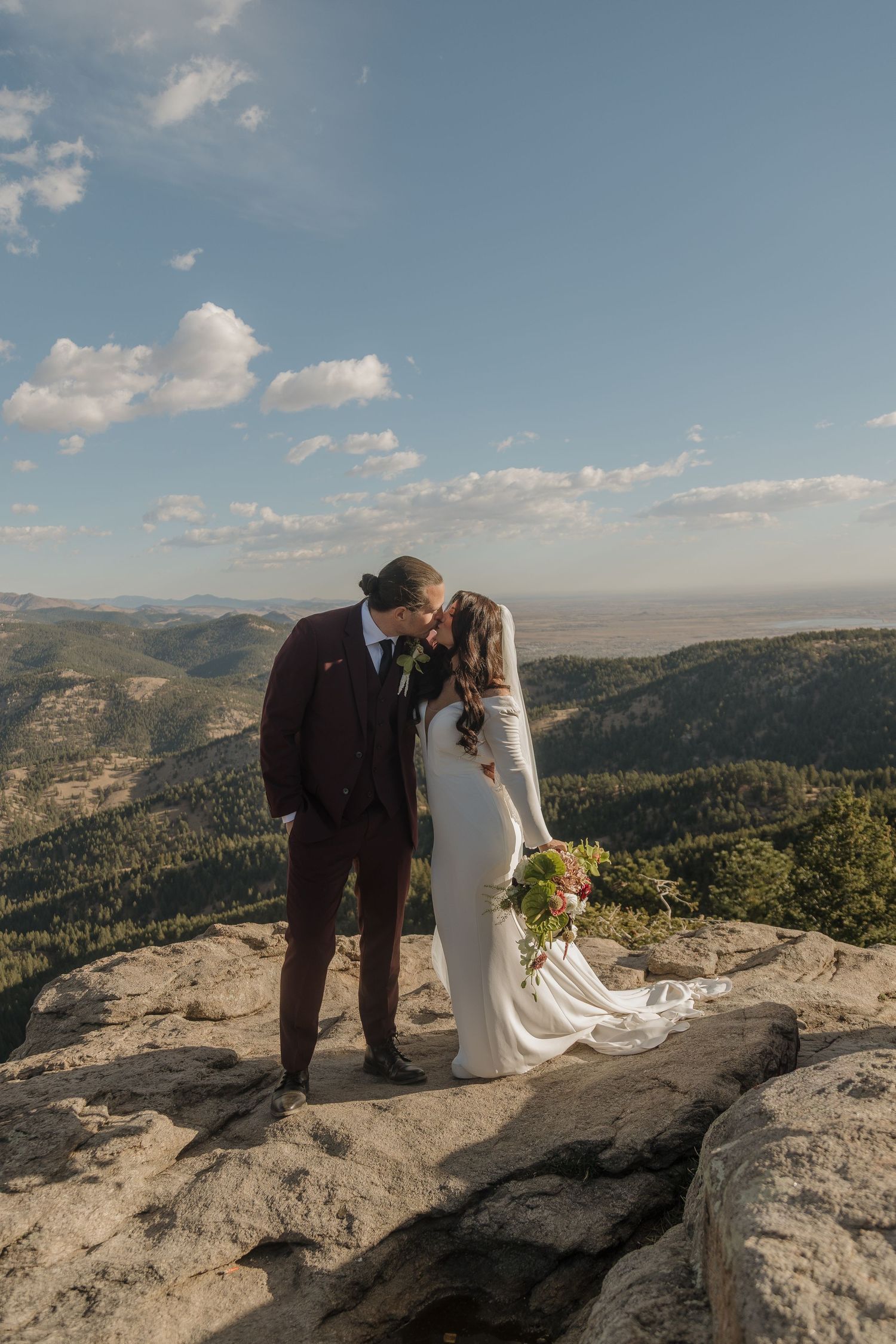 Bride and groom share a kiss on a rocky mountain summit with sweeping valley views on their wedding day.