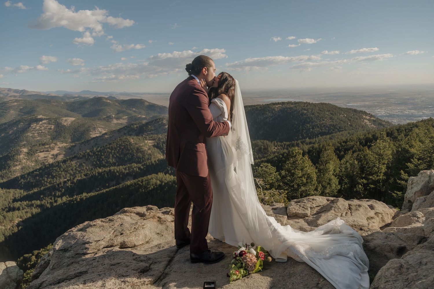 Bride and groom share a kiss on mountain summit with panoramic valley views behind them.