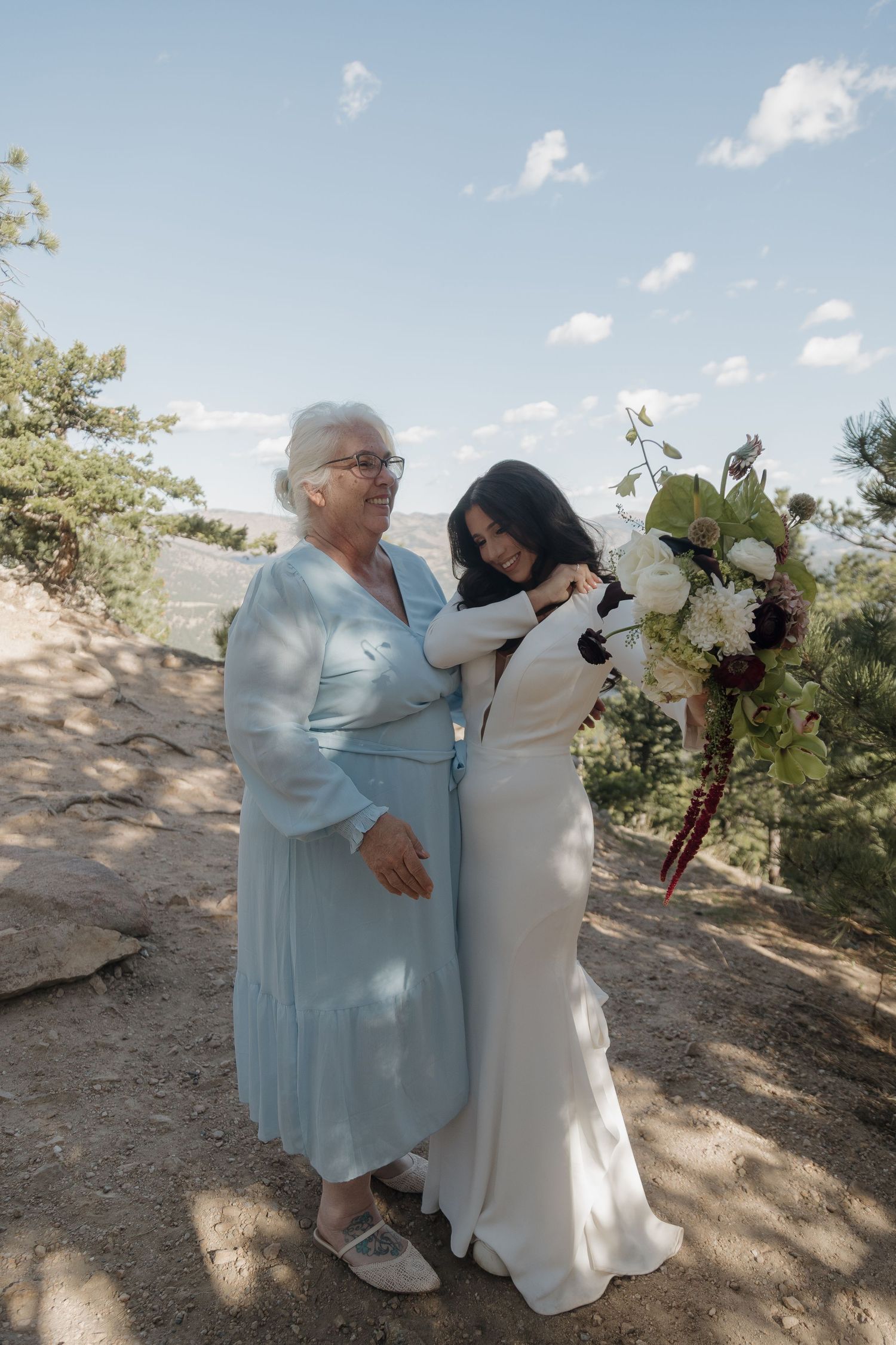 Bride in white gown embracing elderly woman in blue dress at outdoor mountain wedding.