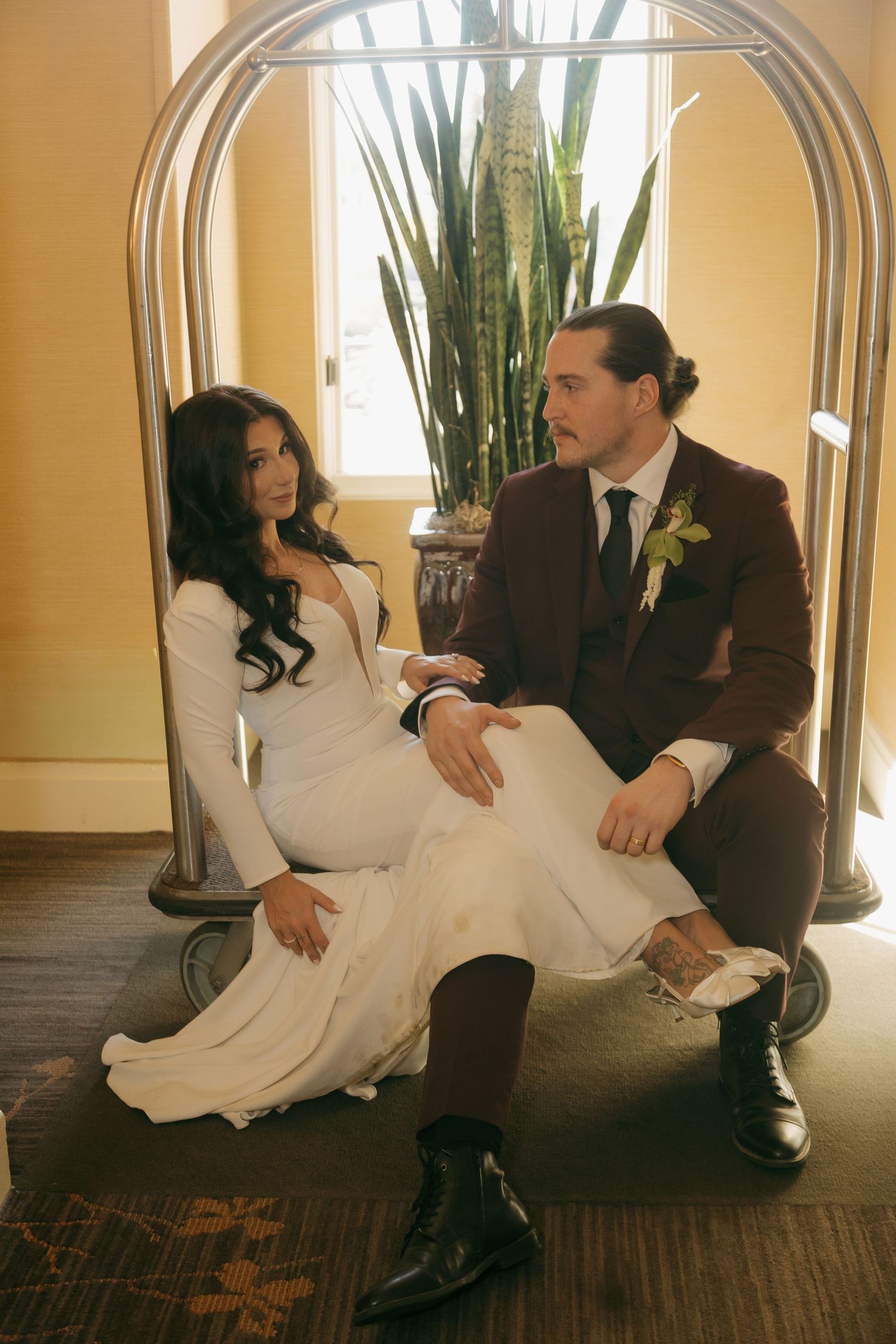 Bride in white long-sleeve gown and groom in burgundy suit sitting in hotel luggage cart.