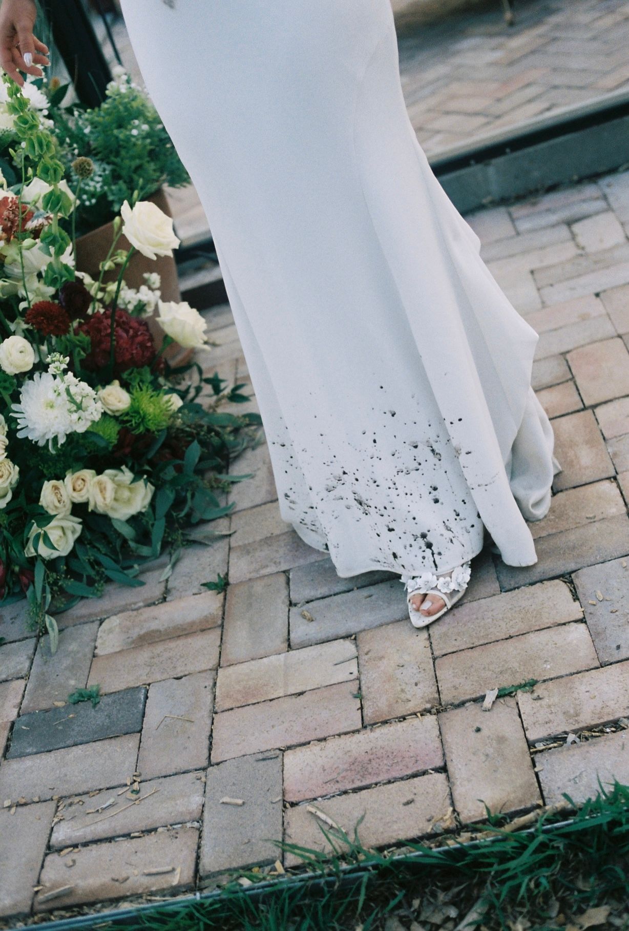 Close-up of a brides white wedding gown hem with dirt specks beside a lush floral bouquet on brick pavers.